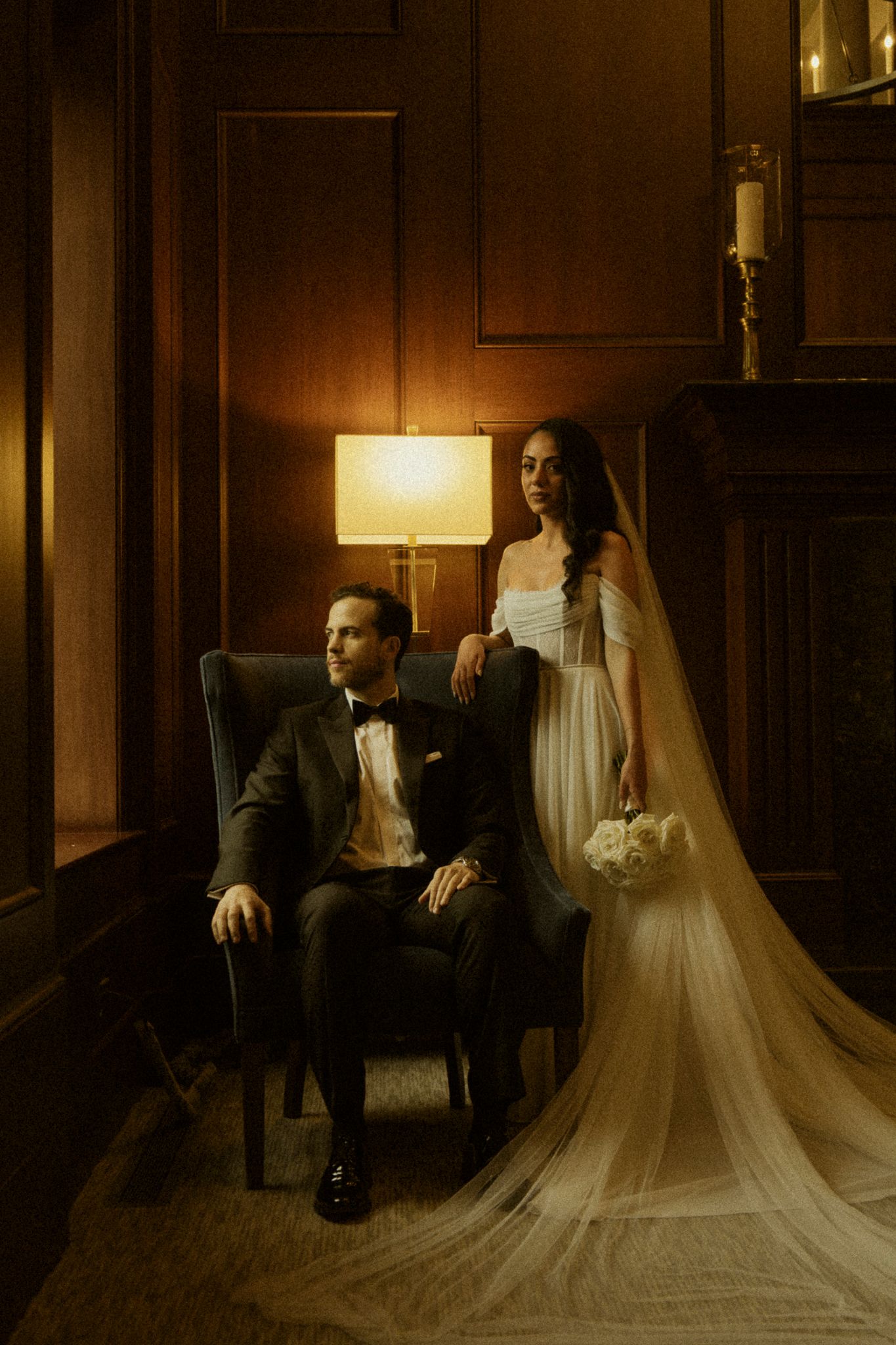 Elegant bride in white gown with veil holds bouquet beside groom in tuxedo seated in armchair in moody lit room.