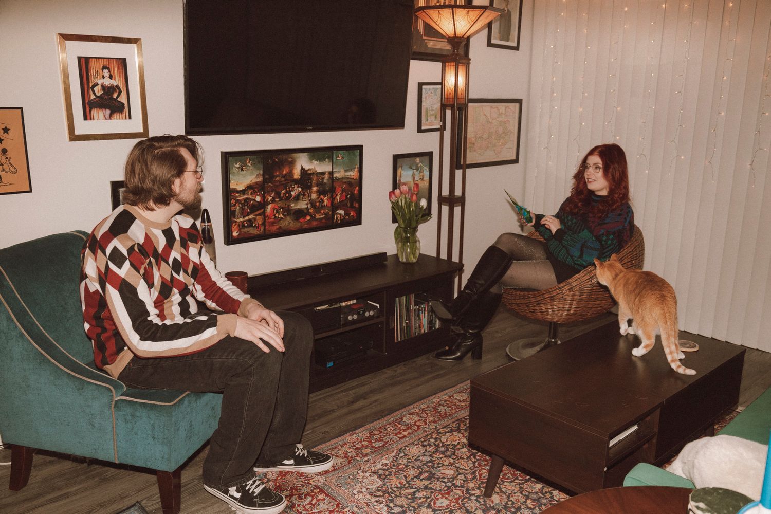 Two people watching TV in a cozy living room with a cat on the couch and vintage decor on the walls.