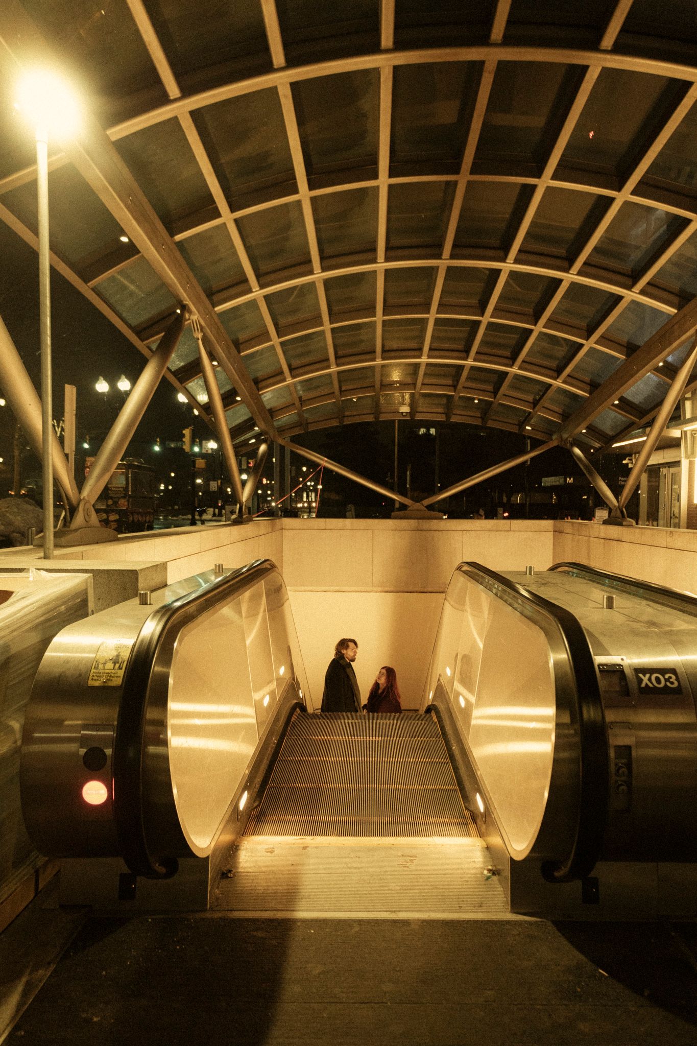 Modern subway station interior with curved glass ceiling and escalators illuminated in warm lighting at night.