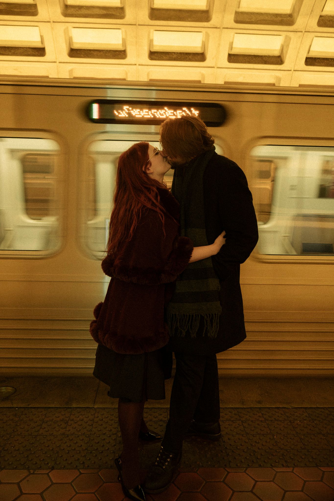 A couple embraces in front of a moving subway train at a station with hexagonal floor tiles.