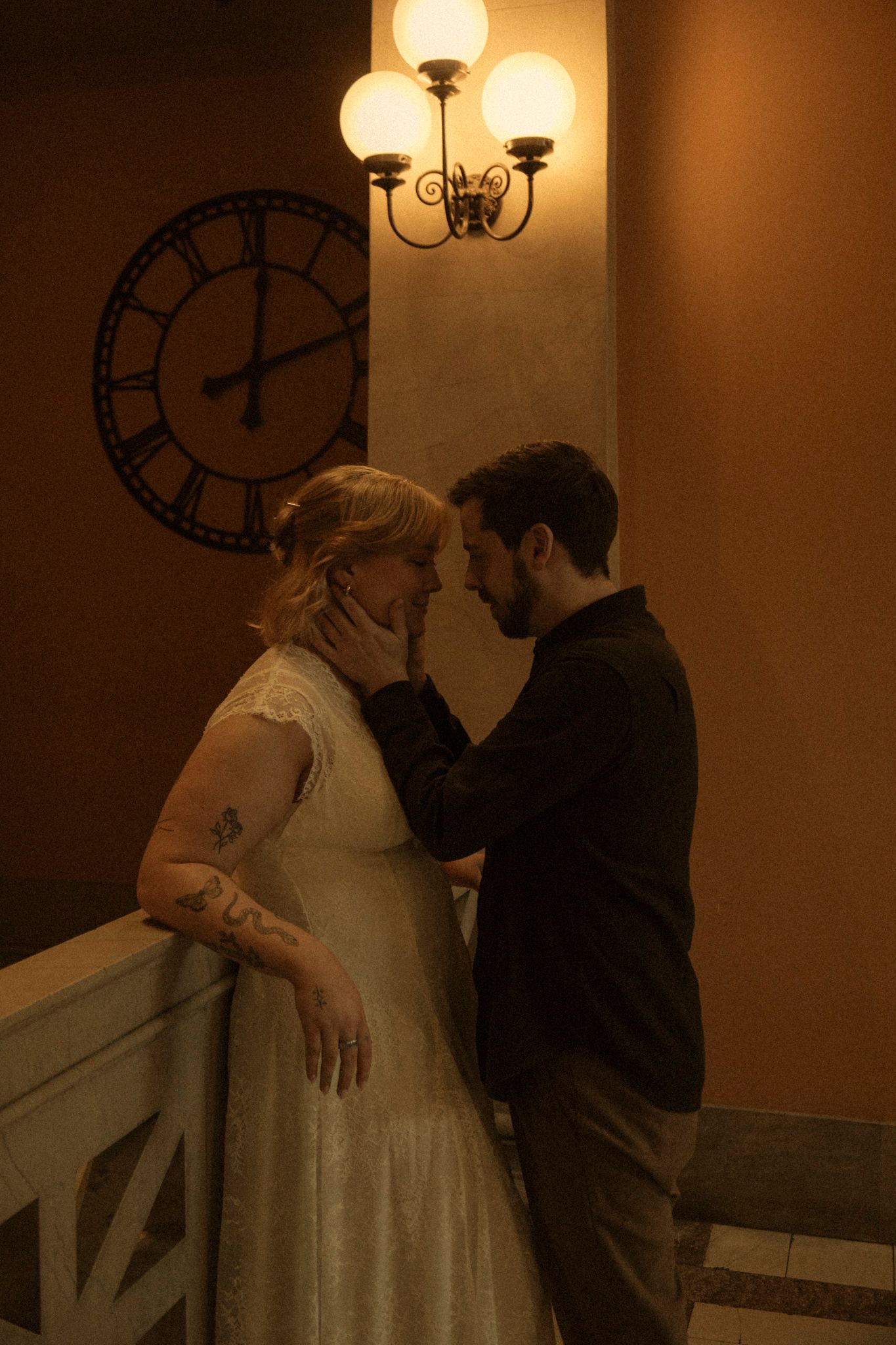 A romantic couple stands intimately in a dimly lit hallway with a vintage wall clock and antique lamp fixture above.