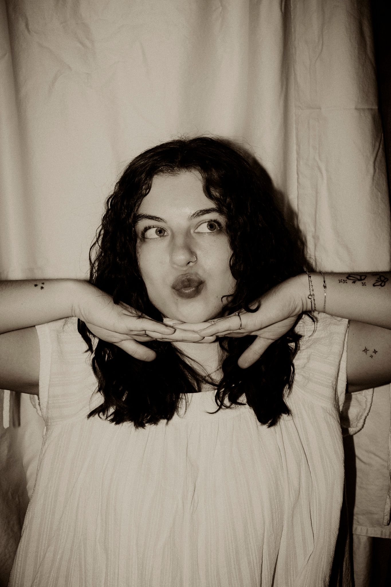 Sepia-toned portrait of a young woman with dark curly hair posing with hands crossed under chin near white curtain.