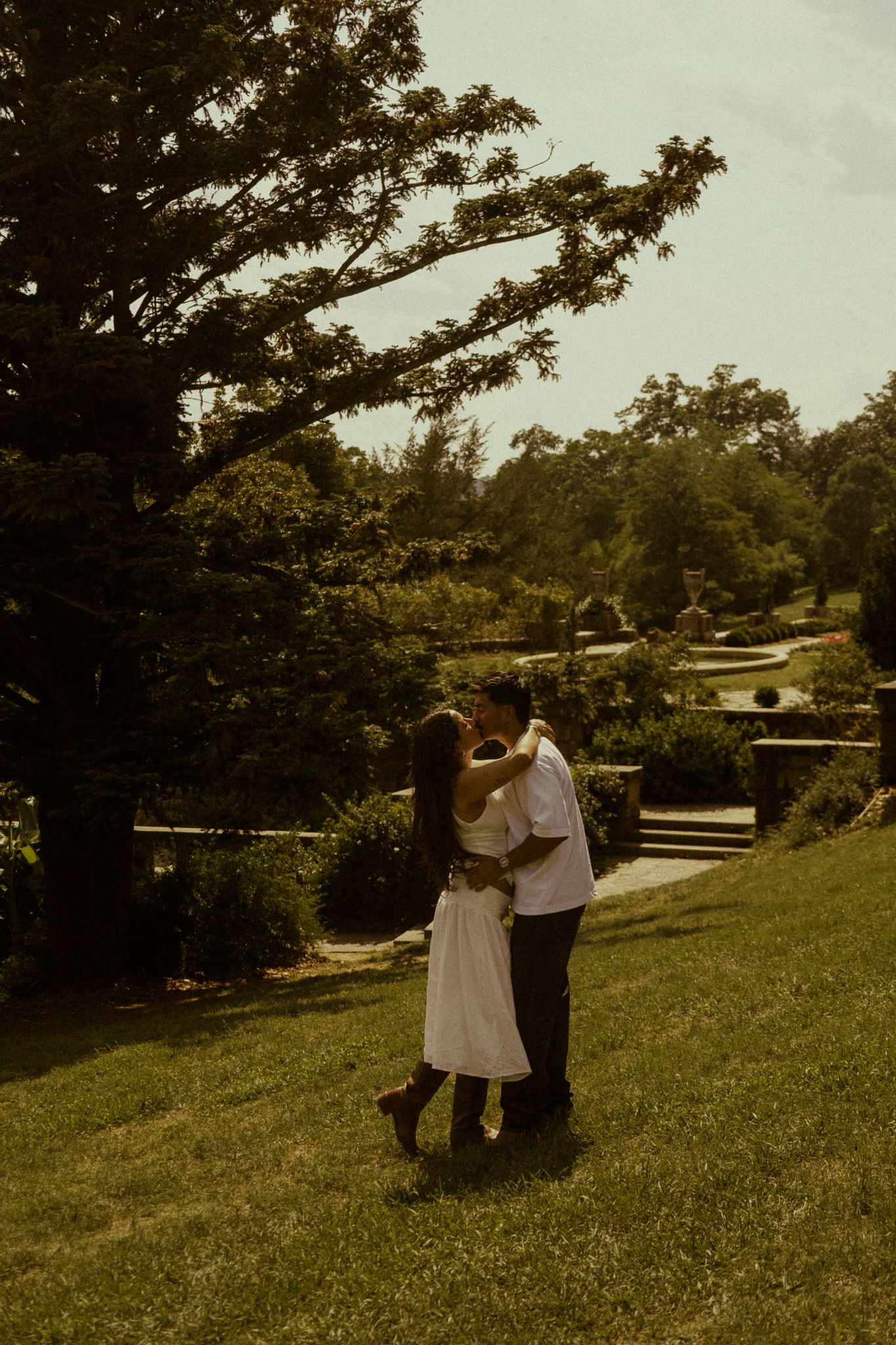 Couple sharing a romantic kiss on a lush green lawn surrounded by trees and gardens on a sunny day.