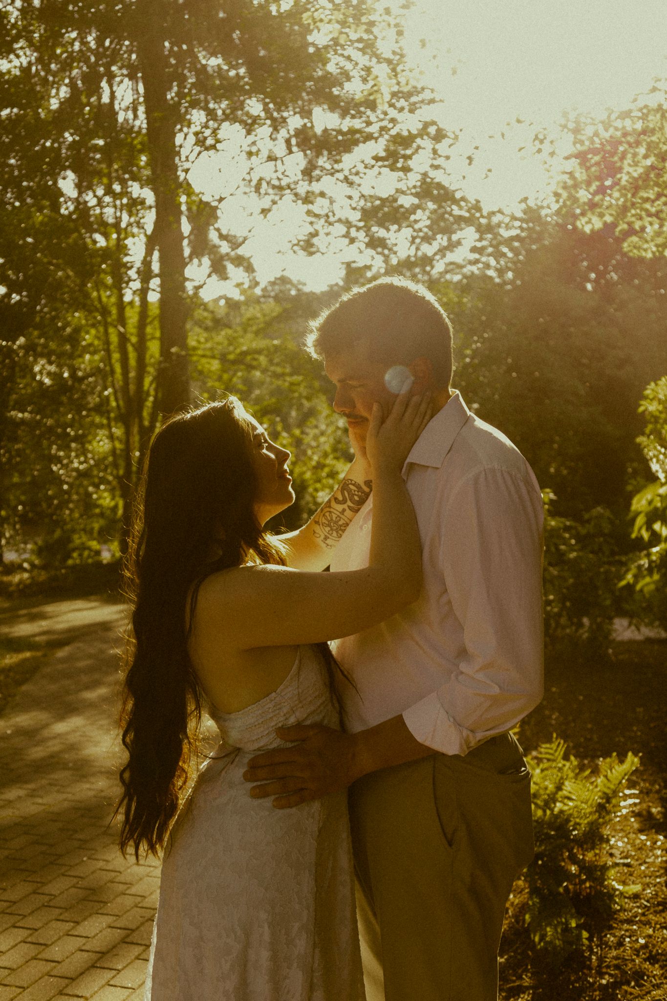 Couple embracing in golden sunset light outdoors, bride in lace dress touching groom's face, surrounded by lush green trees.