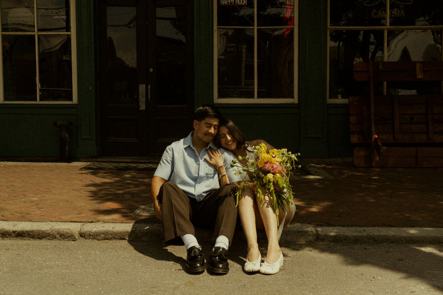 Couple sitting together on a curb holding a colorful bouquet of flowers outside a green storefront.