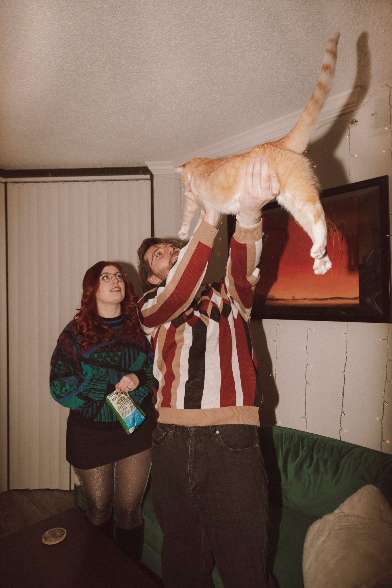 Two people in casual attire playfully lift an orange and white cat up in the air in a living room setting.
