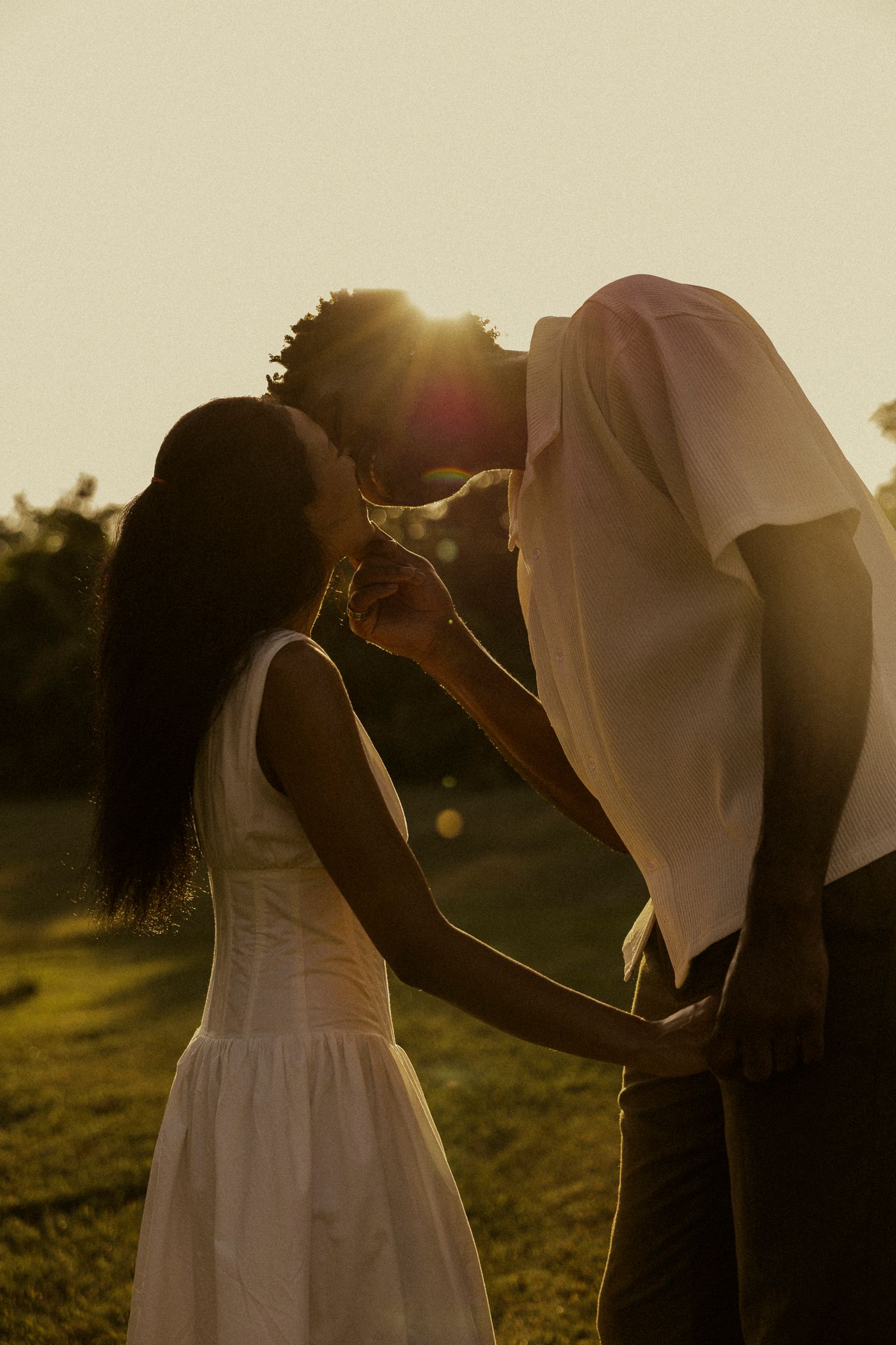 Couple sharing a romantic kiss at golden hour sunset in a green field, woman in white dress.