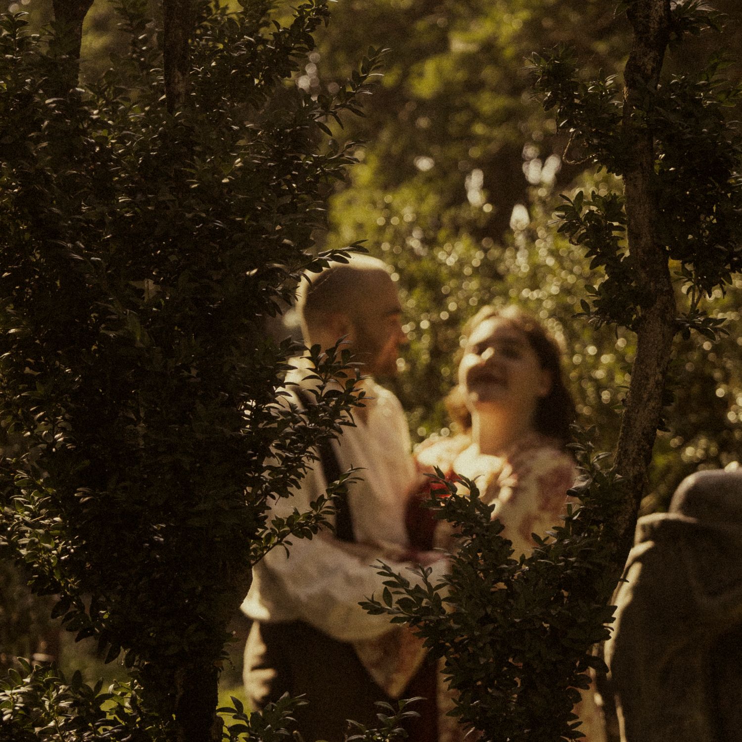 A couple shares a romantic moment surrounded by lush green foliage in a sunlit garden setting.