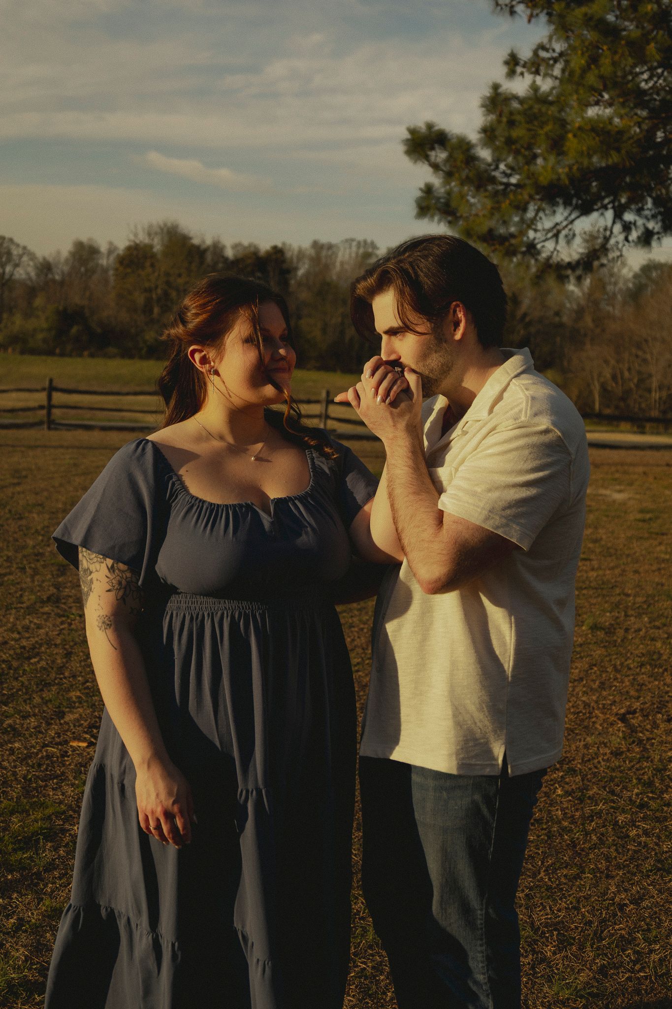 A man in a white shirt kisses a woman's hand outdoors at golden hour near a wooden fence.