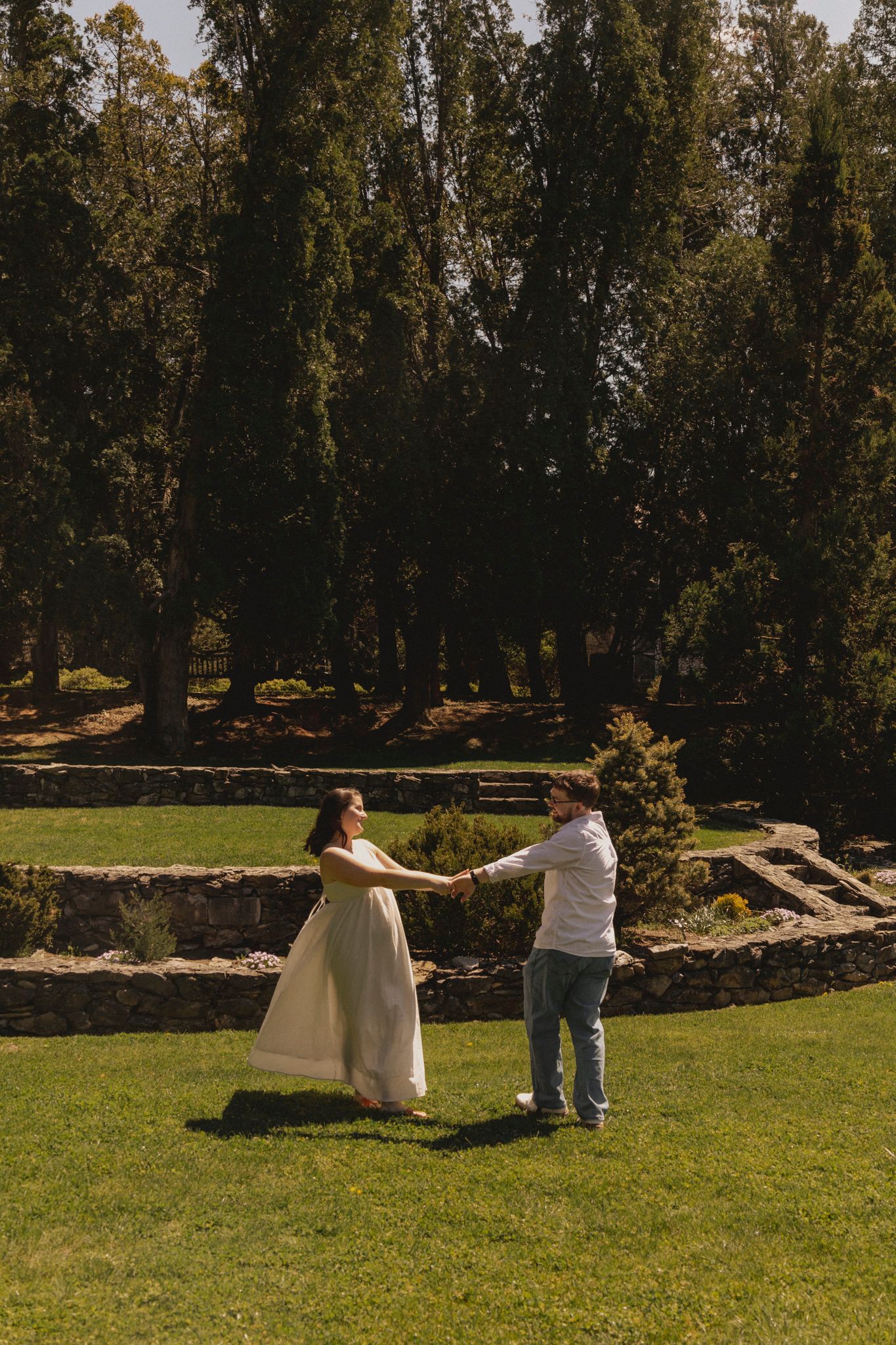Couple holding hands and dancing on green lawn in sunlit garden with stone wall and tall cypress trees backdrop.
