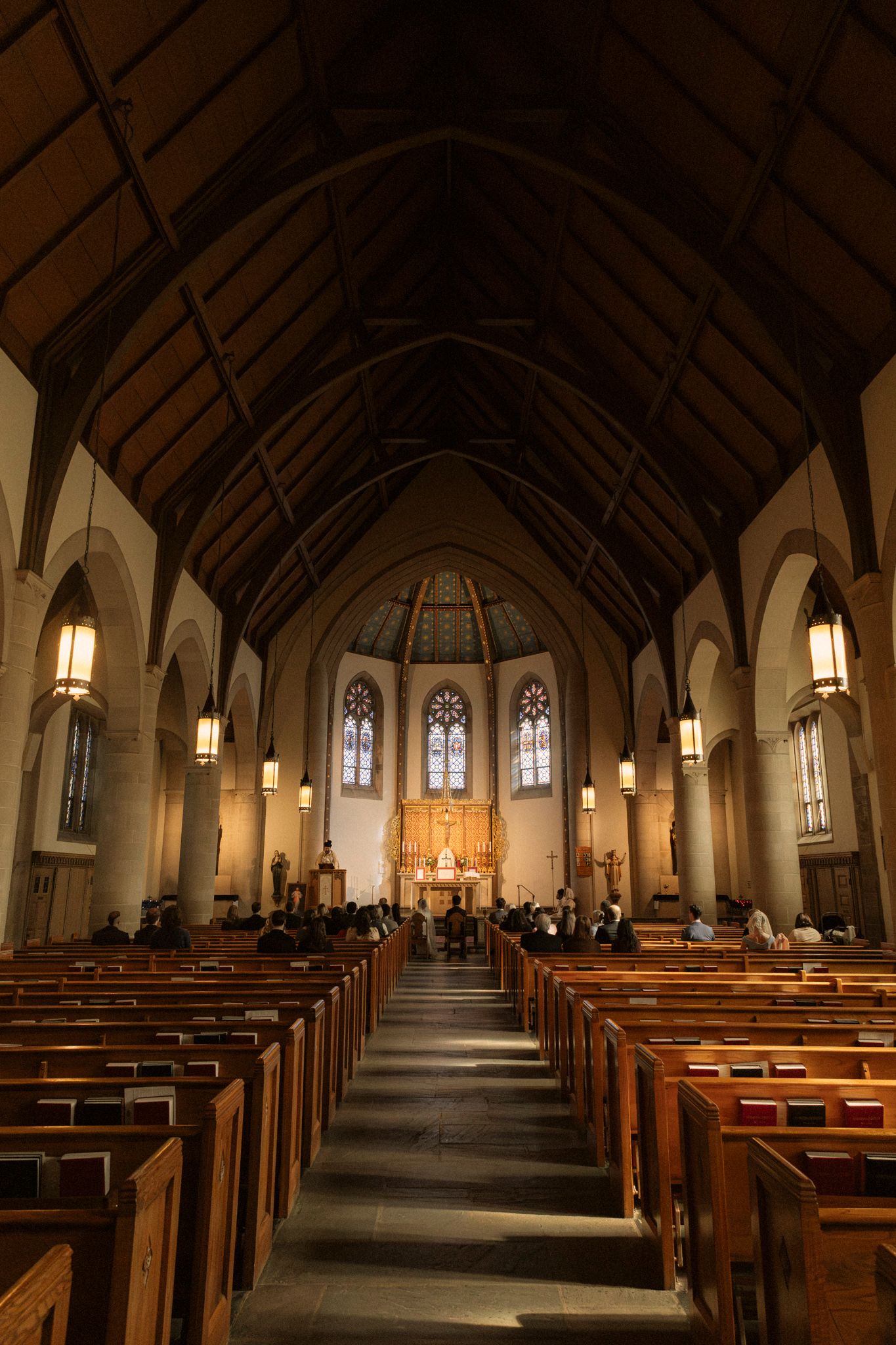 Gothic church interior with wooden pews leading to ornate altar, vaulted ceiling, and stained glass windows during a wedding ceremony.