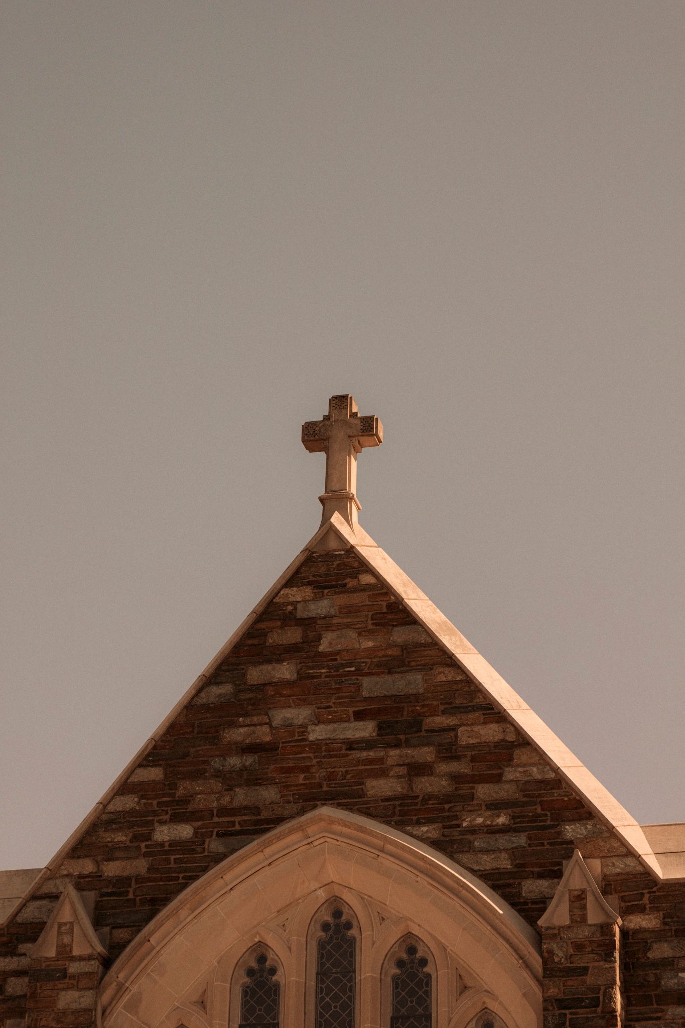 Stone church gable with a cross atop, featuring Gothic arched windows and brick facade under a clear sky.