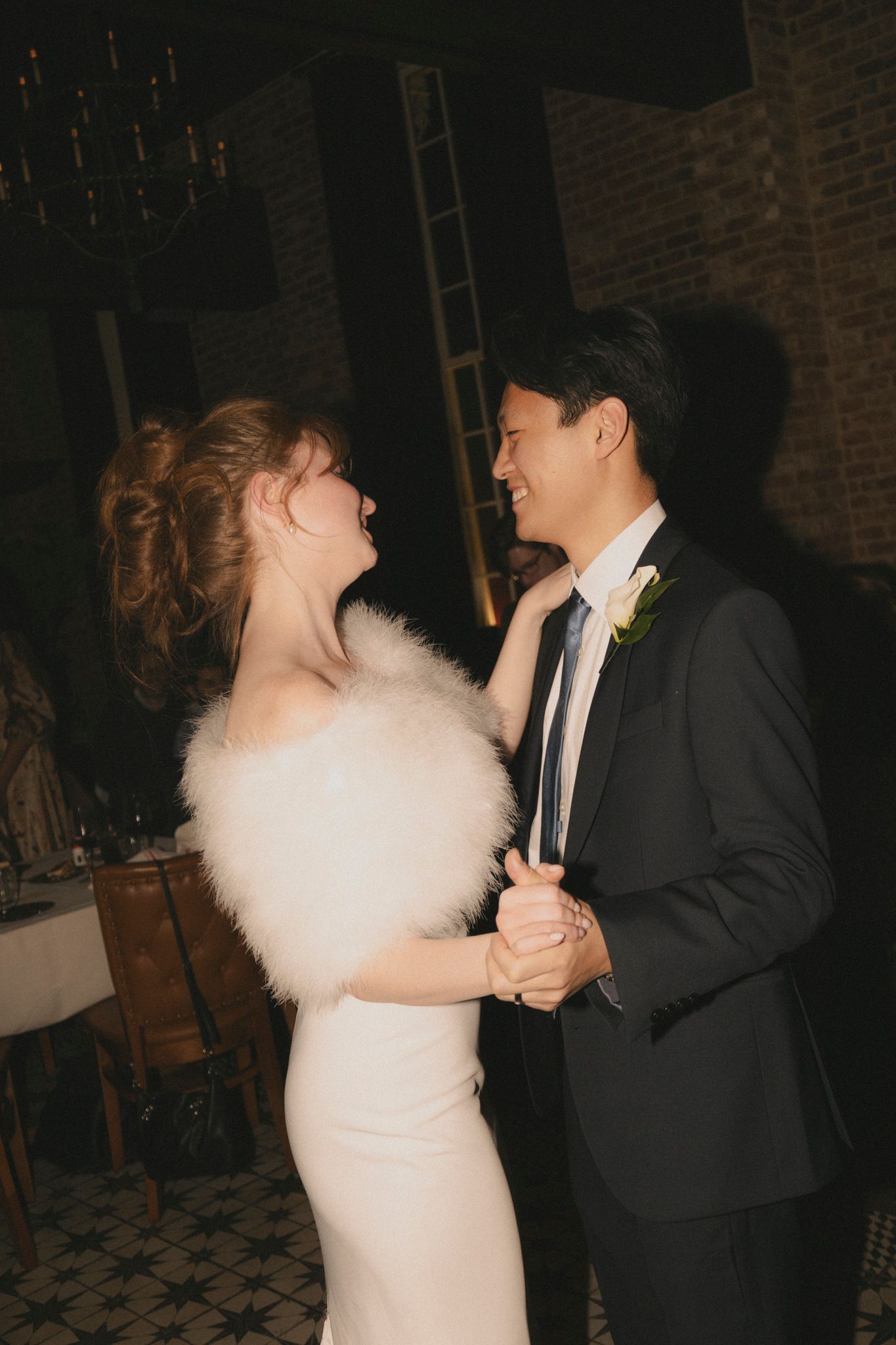 Bride and groom share first dance, she wears white gown with feather wrap, he wears dark suit with boutonniere.