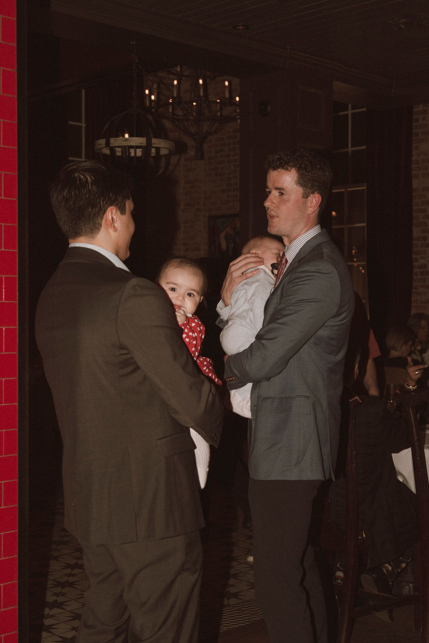 Two men in gray suits stand with a child and baby at a formal indoor event with chandelier lighting.