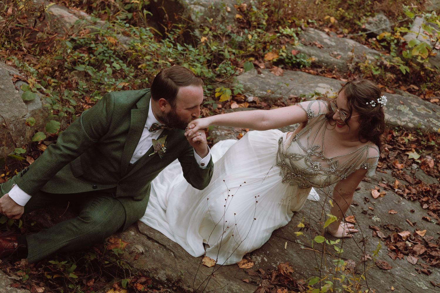 A couple in wedding attire playfully poses together on autumn forest ground covered in fallen leaves.
