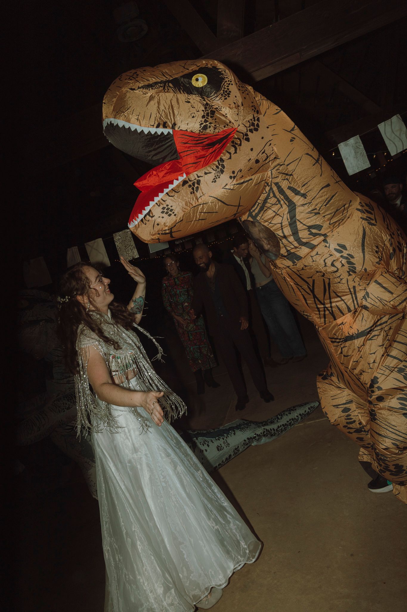 A person in a white dress dances with someone wearing an inflatable T-Rex dinosaur costume at a party.