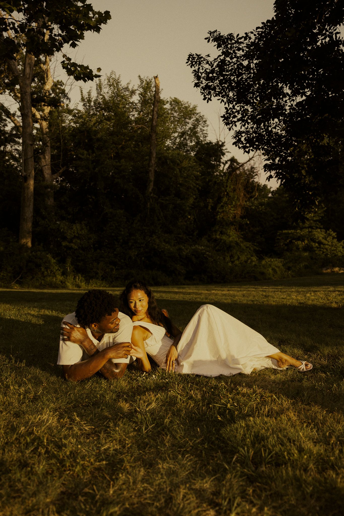 Couple lying on grass at golden hour, woman in white dress, surrounded by lush green trees in a park.