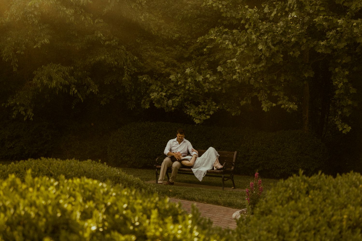 Couple relaxing on a park bench surrounded by lush green hedges and trees in warm golden sunlight.