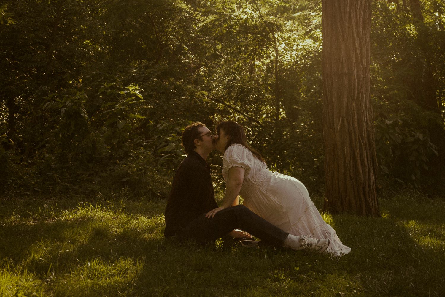 Couple sharing a romantic kiss while sitting on lush green grass beneath a sunlit tree in a serene forest setting.