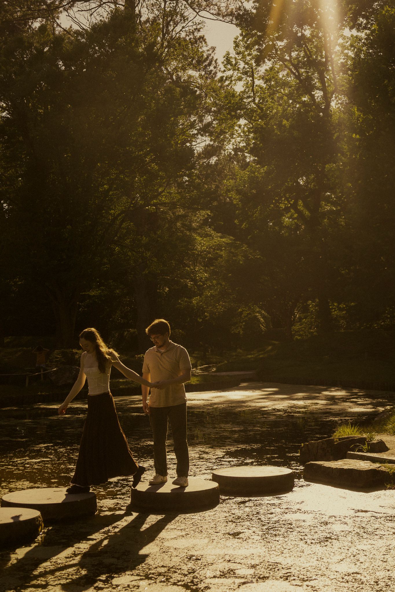 Couple holds hands walking on stepping stones in golden sunlight filtering through lush green trees in a forest.