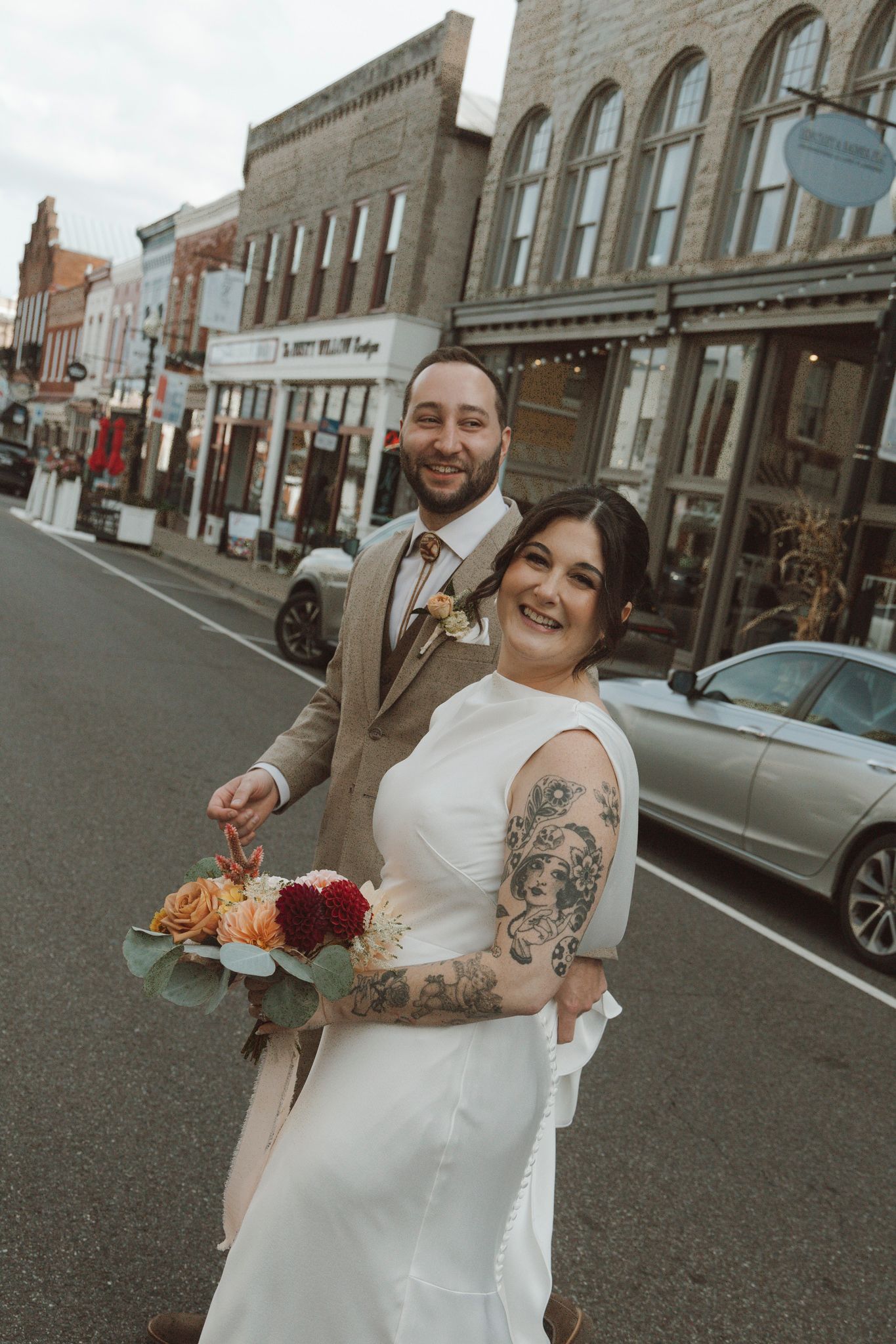 A joyful wedding couple poses on a city street with brick buildings in the background.