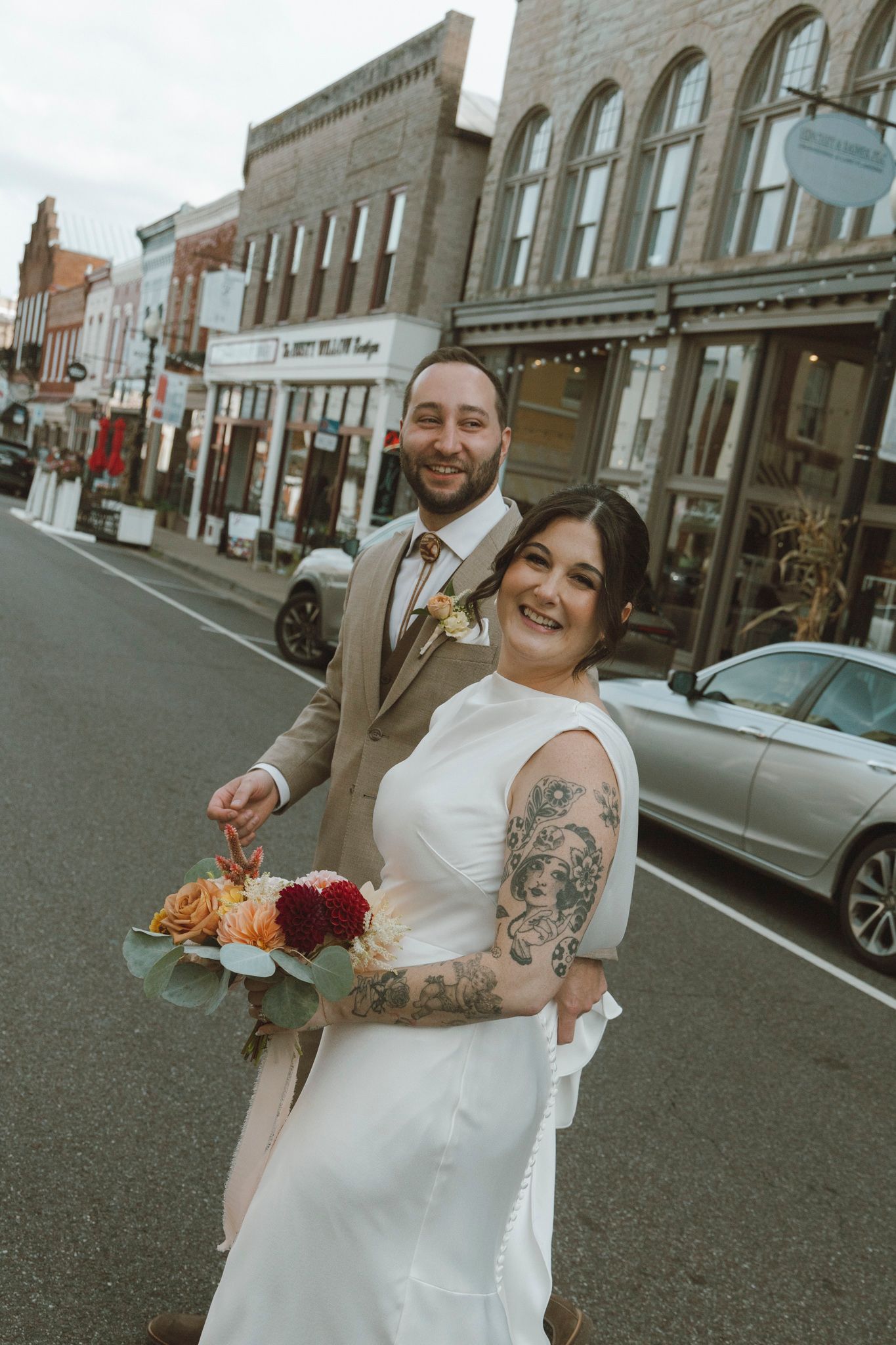 A couple in wedding attire share a joyful moment together on a city street with historic brick buildings in the background.