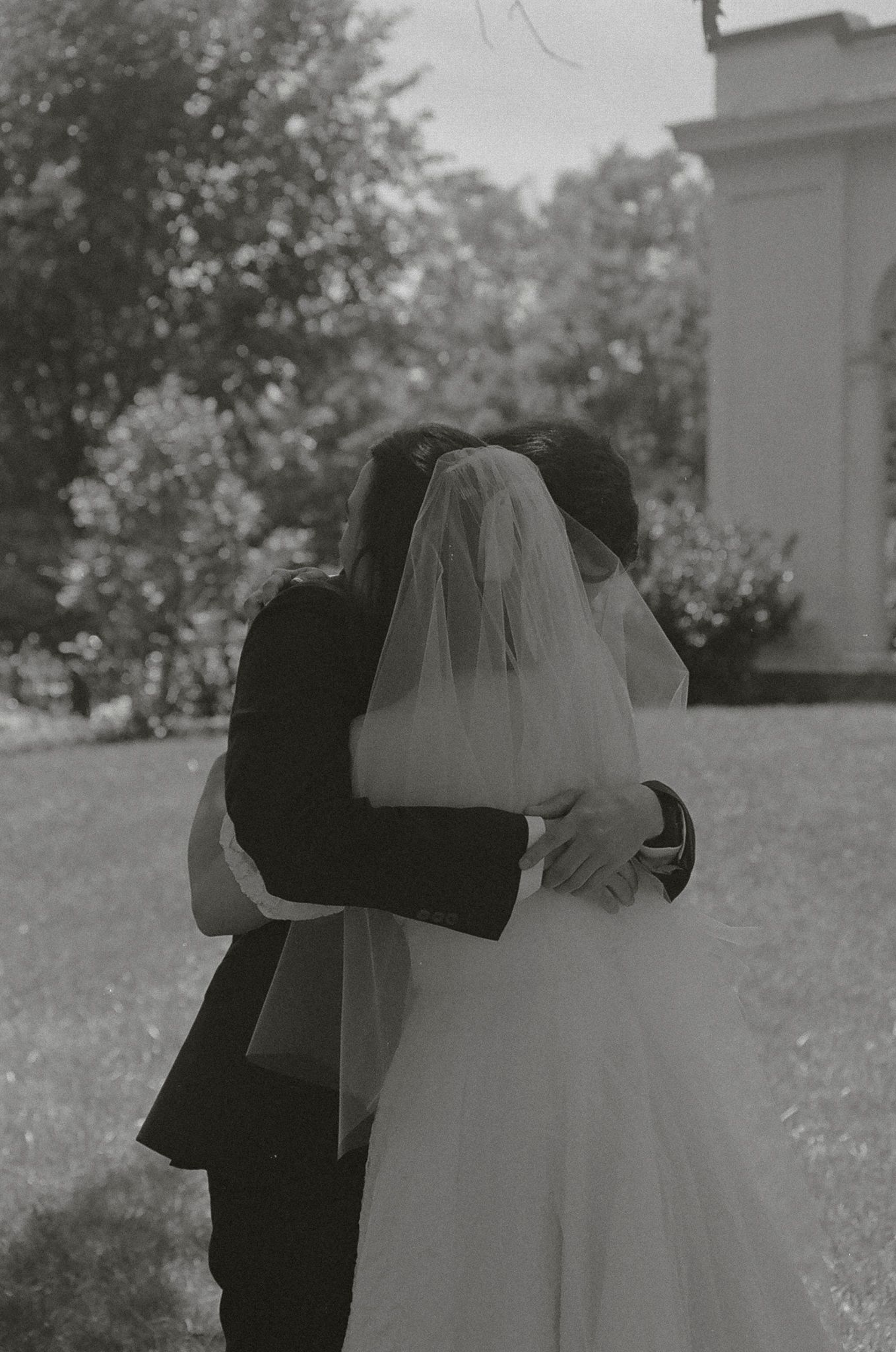A black and white wedding photograph showing a couple embracing outdoors, with the bride in a white gown and veil.