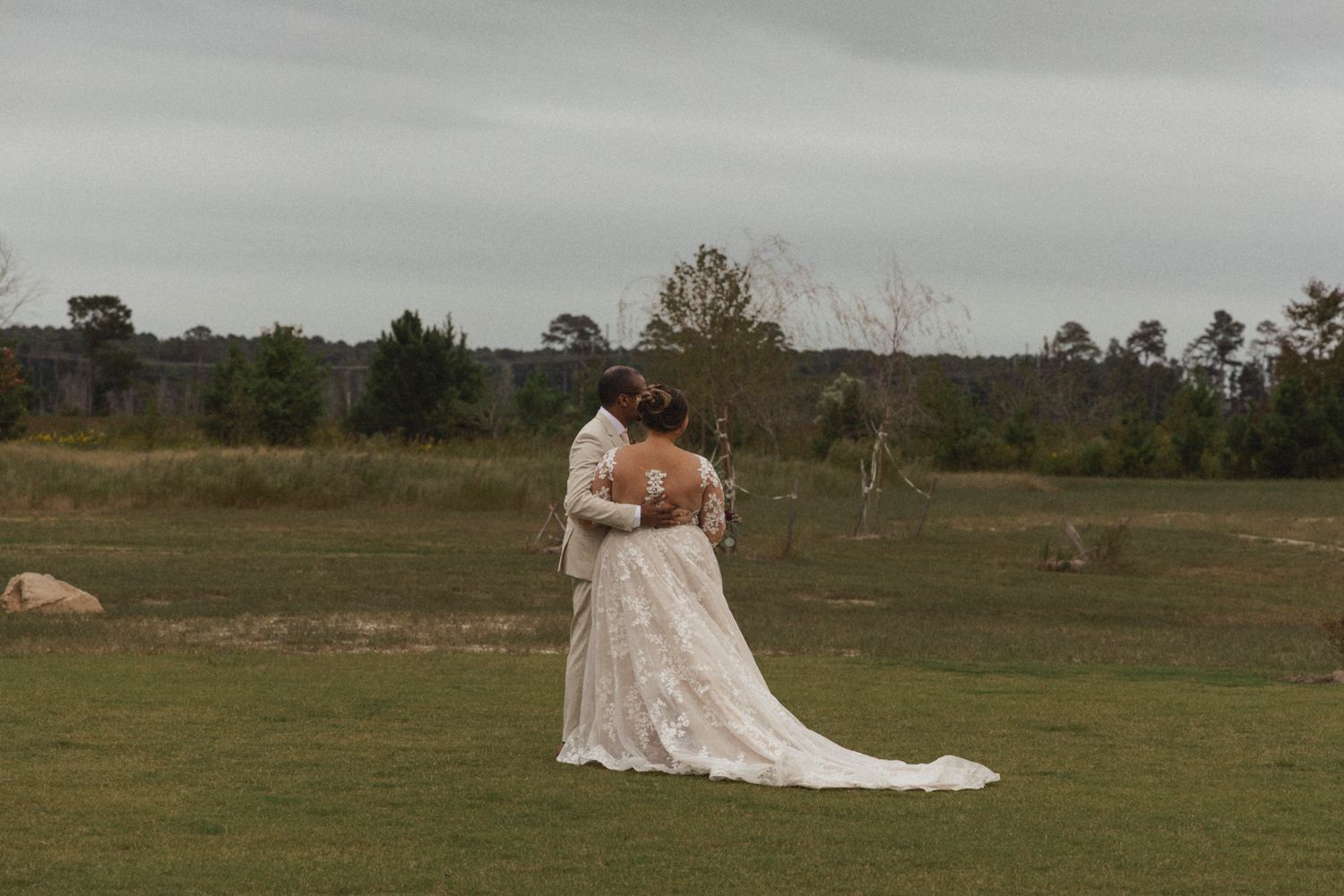A romantic couple poses in a grassy field wearing elegant wedding attire with a long flowing white dress.