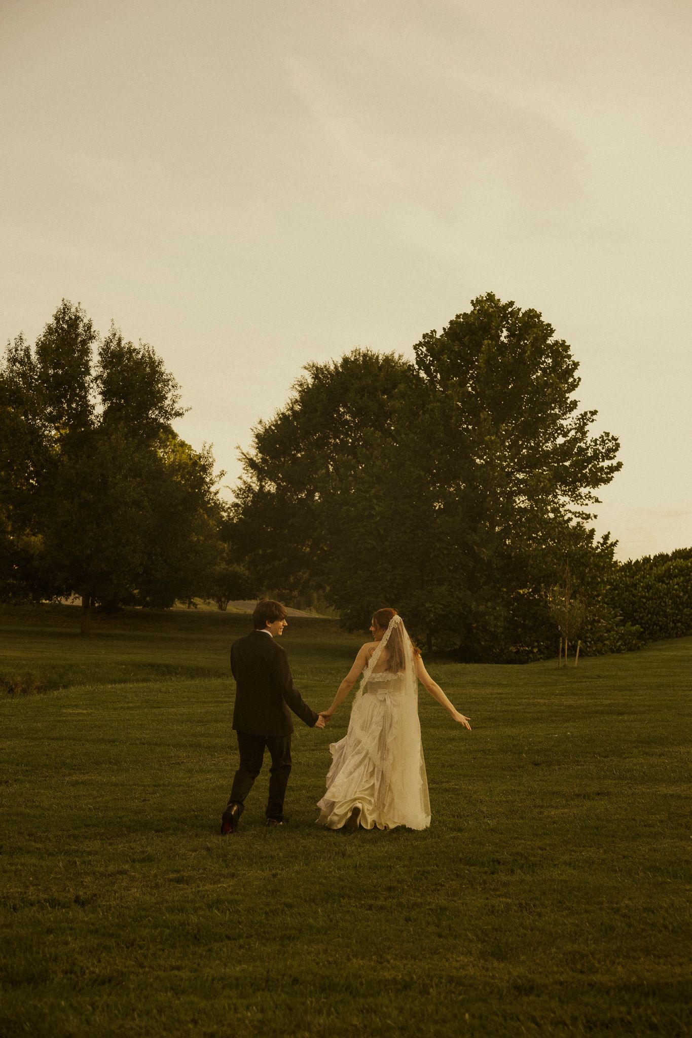 A bride and groom hold hands walking across a lush green lawn at dusk with trees in the background.