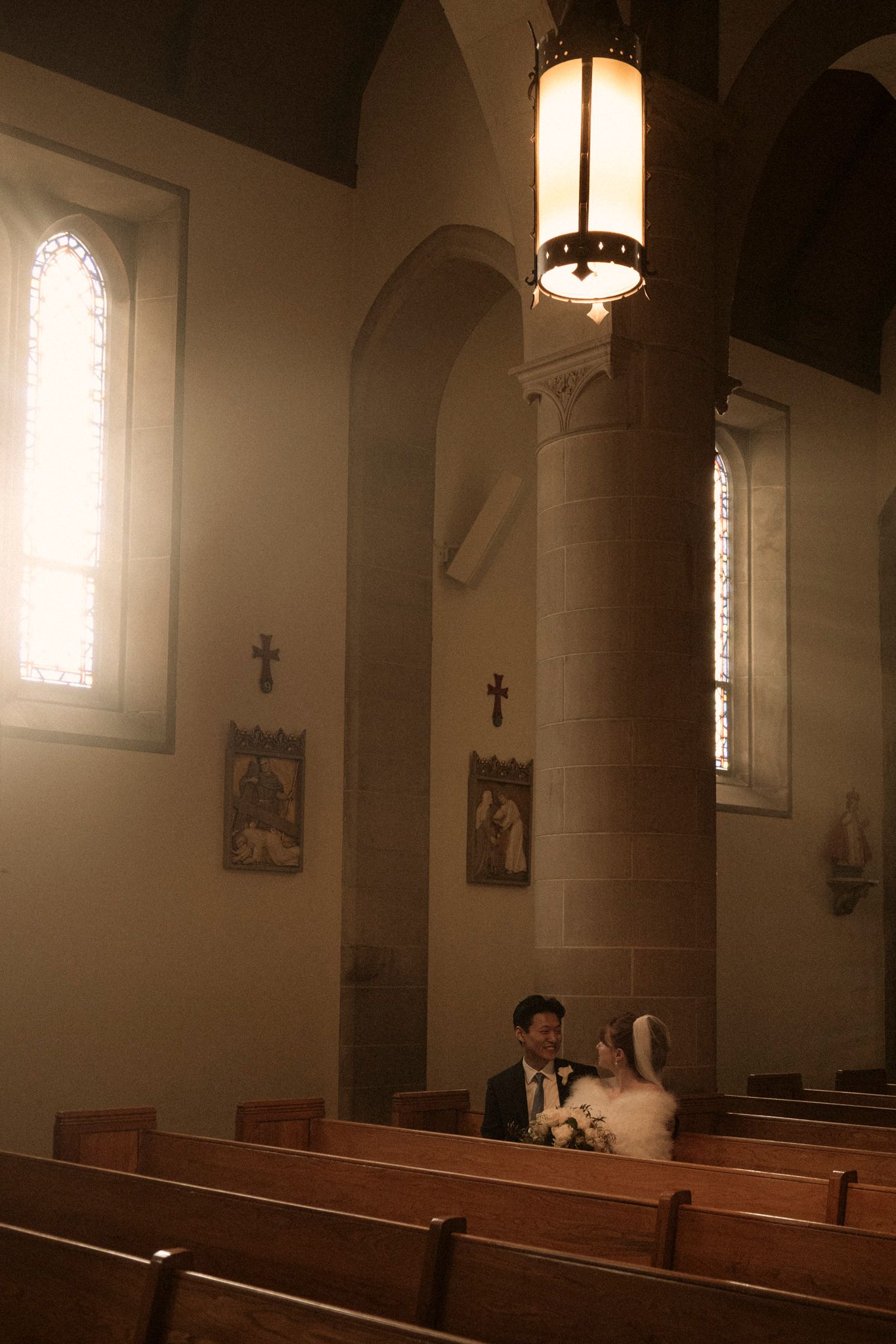 Bride and groom share an intimate moment in a warmly lit Catholic church with wooden pews and stone arches.