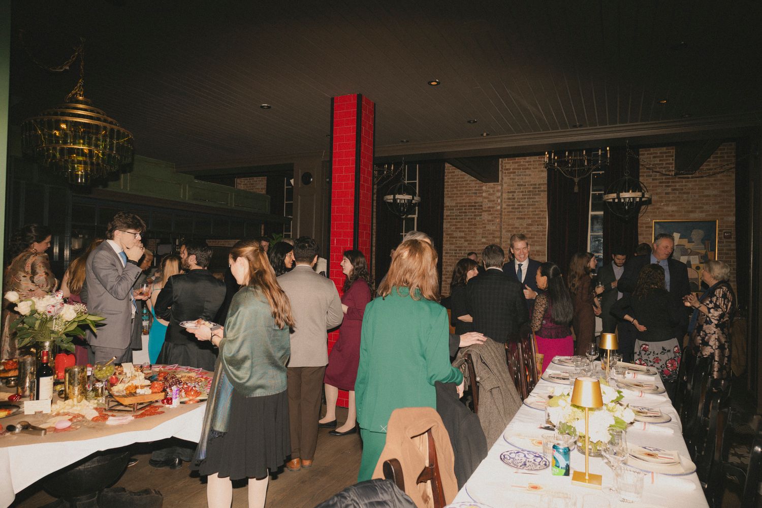 Guests mingle at an elegant indoor dinner party with long tables set with food and floral arrangements.