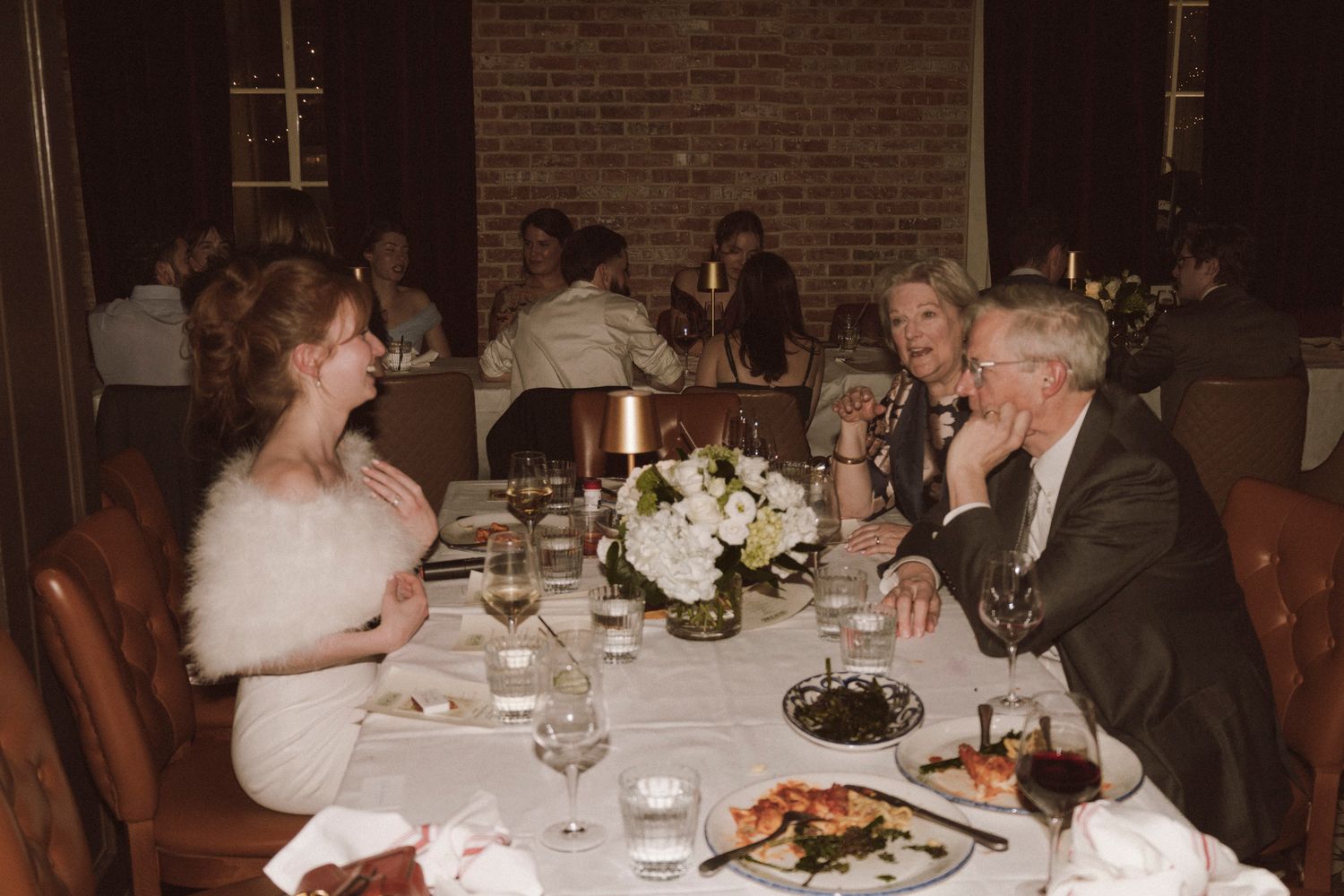 Bride in white fur stole laughing with guests at an elegant wedding reception dinner table.