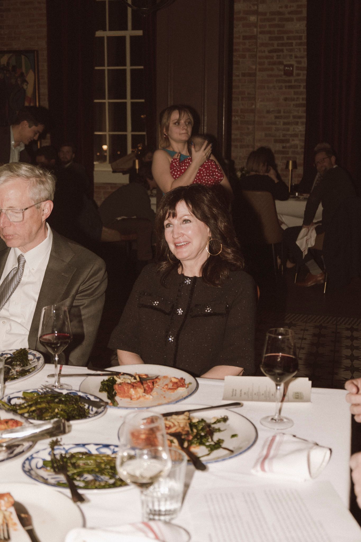 Smiling woman in dark jacket seated at a wedding reception dinner table with wine glasses.