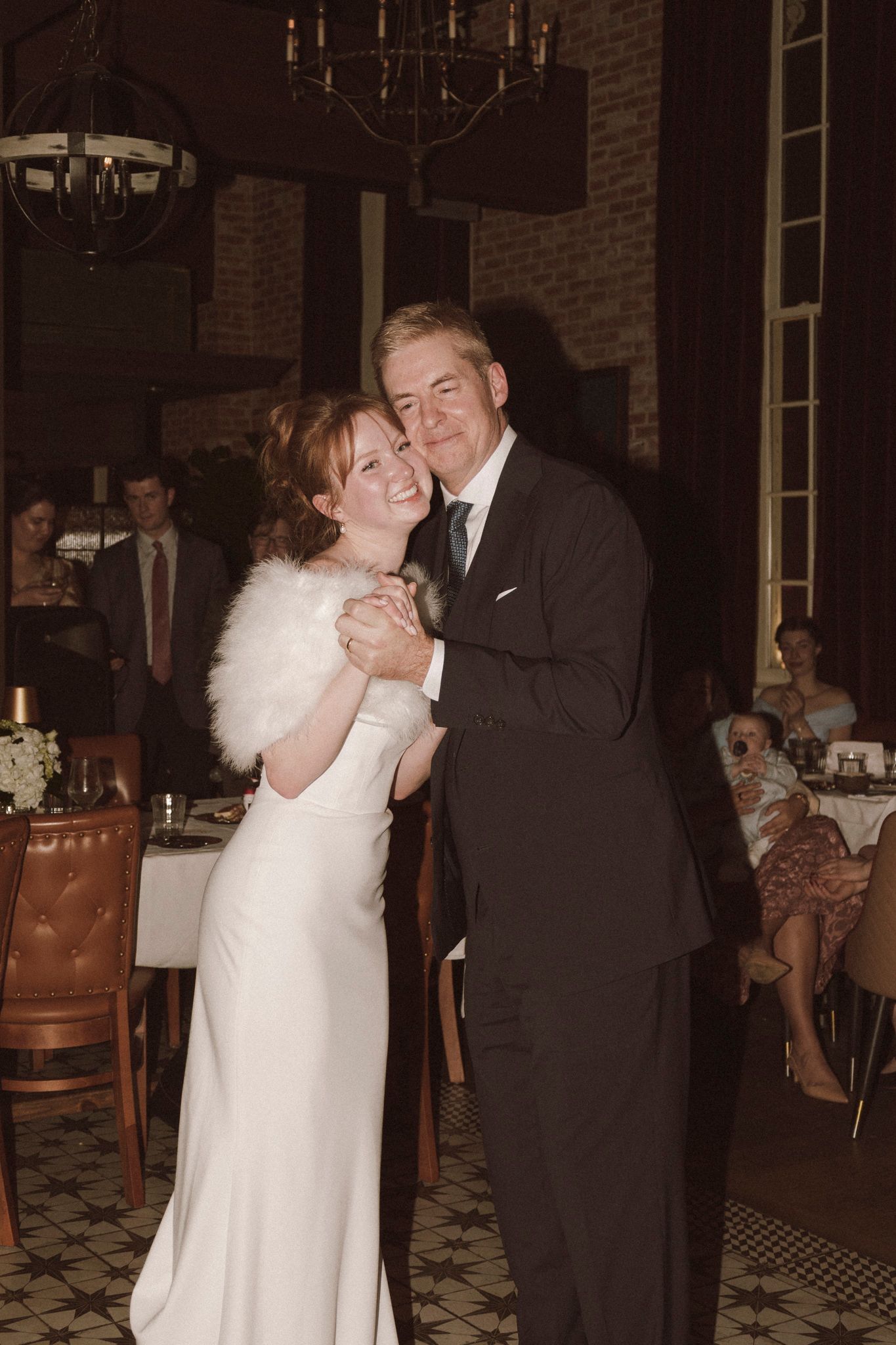 Smiling bride and groom sharing first dance in elegant reception venue with brick walls, chandeliers, and seated guests.