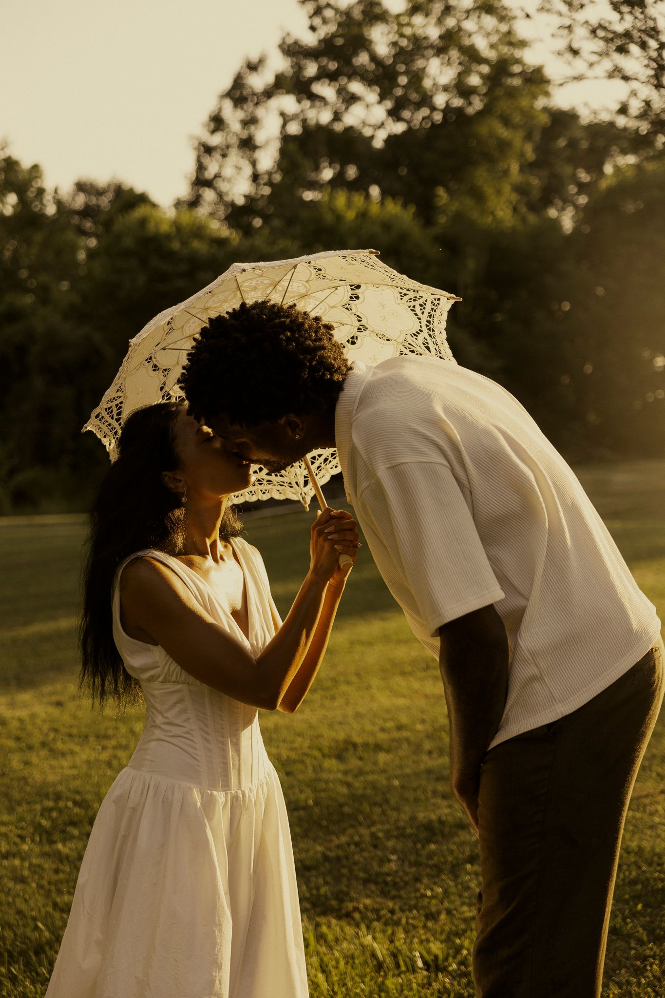 A couple shares a romantic kiss under a lace umbrella in a sunlit park, dressed in white outfits.