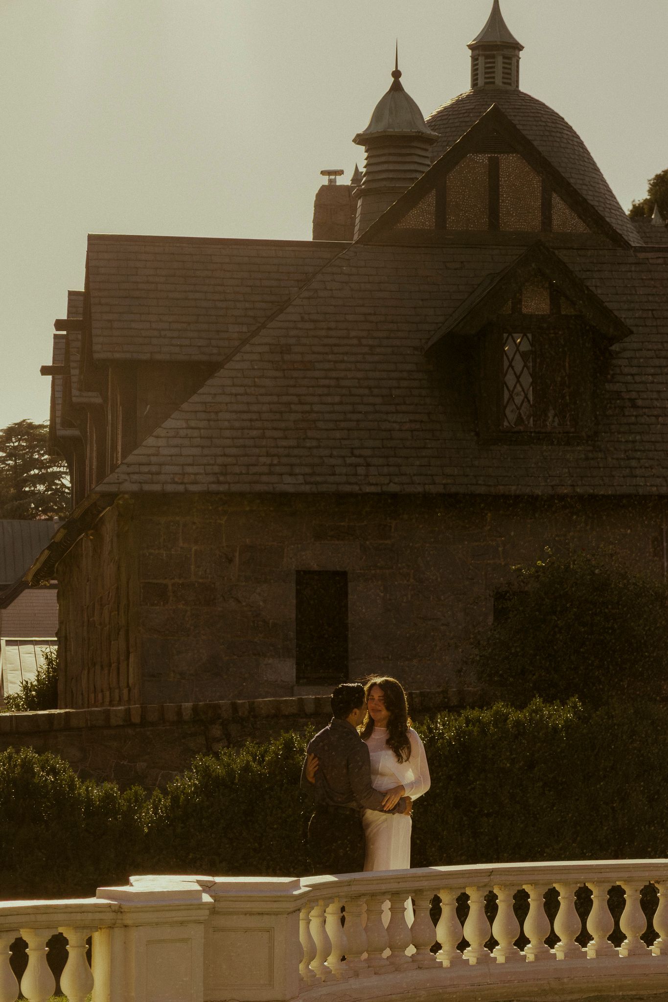 Couple embracing at golden hour in front of a historic stone mansion with ornate Tudor architecture.