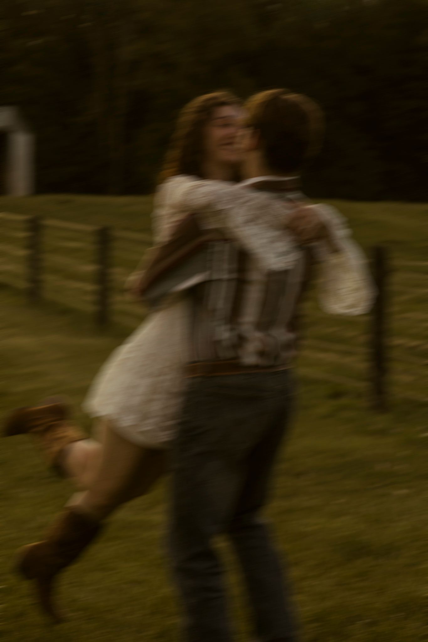 Blurred motion photo of a couple embracing and spinning in a grassy field near a wooden fence at dusk.