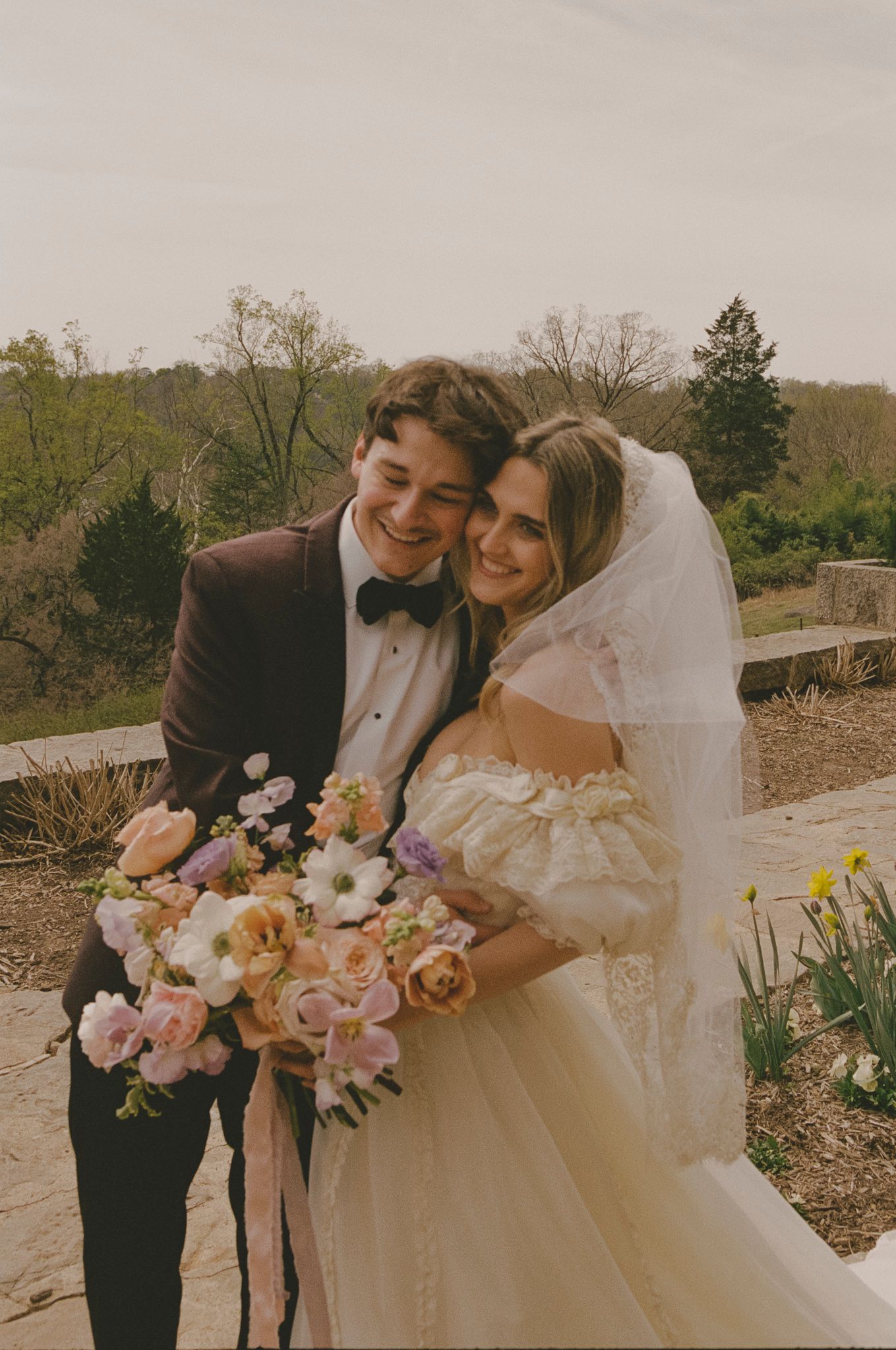 A romantic wedding couple shares intimate moments outdoors while holding a colorful spring bouquet of flowers.