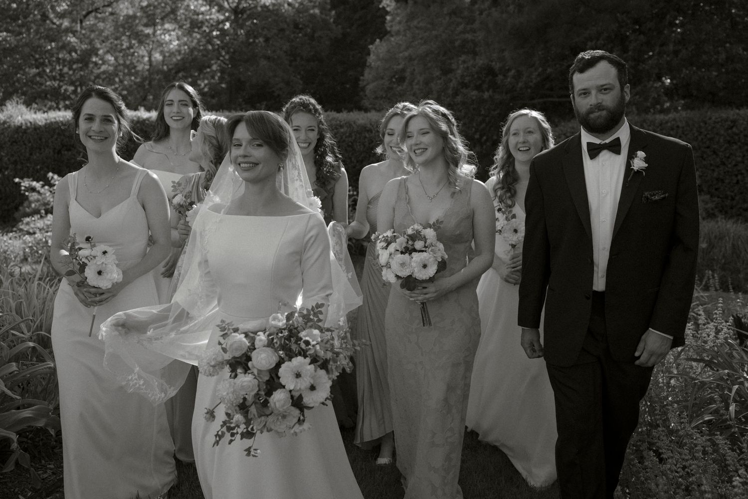 A wedding party poses together outdoors in a garden setting with floral bouquets and formal attire.
