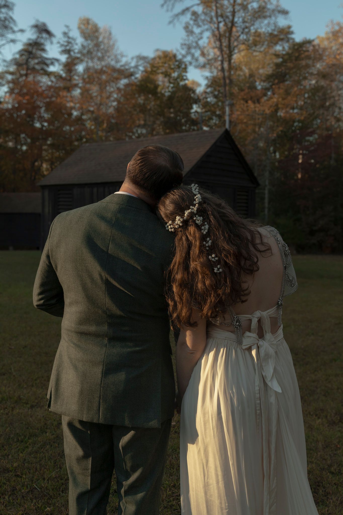 A couple in wedding attire walks together in a field during golden hour with autumn trees in the background.