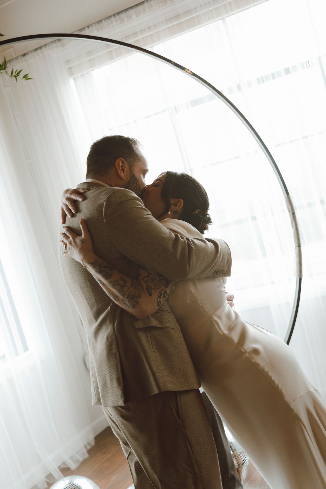 Couple in wedding attire embraces in front of curved arch and sheer curtains with natural lighting.