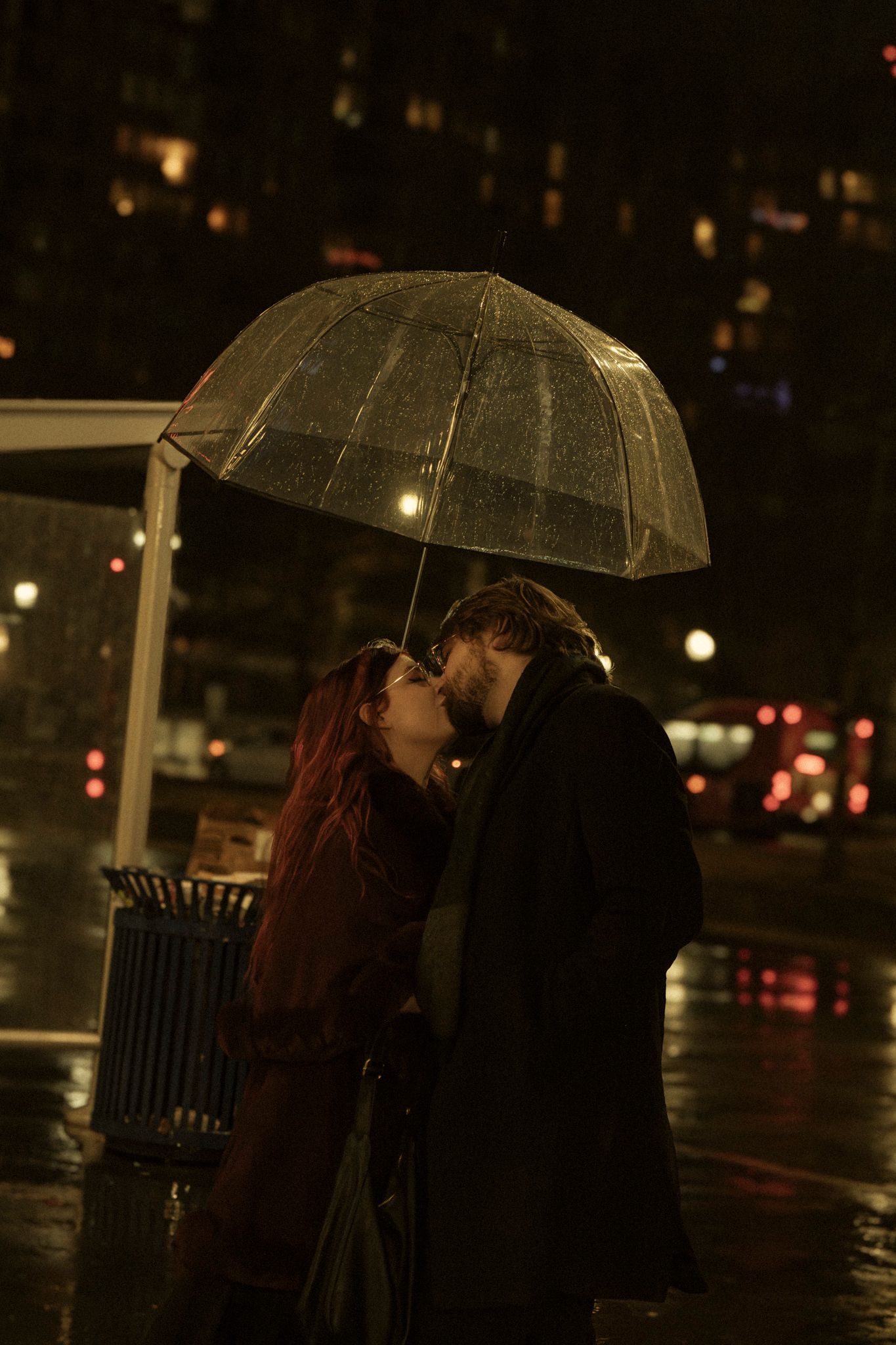 A couple shares an intimate moment under a clear umbrella on a rainy night with city lights twinkling in the background.