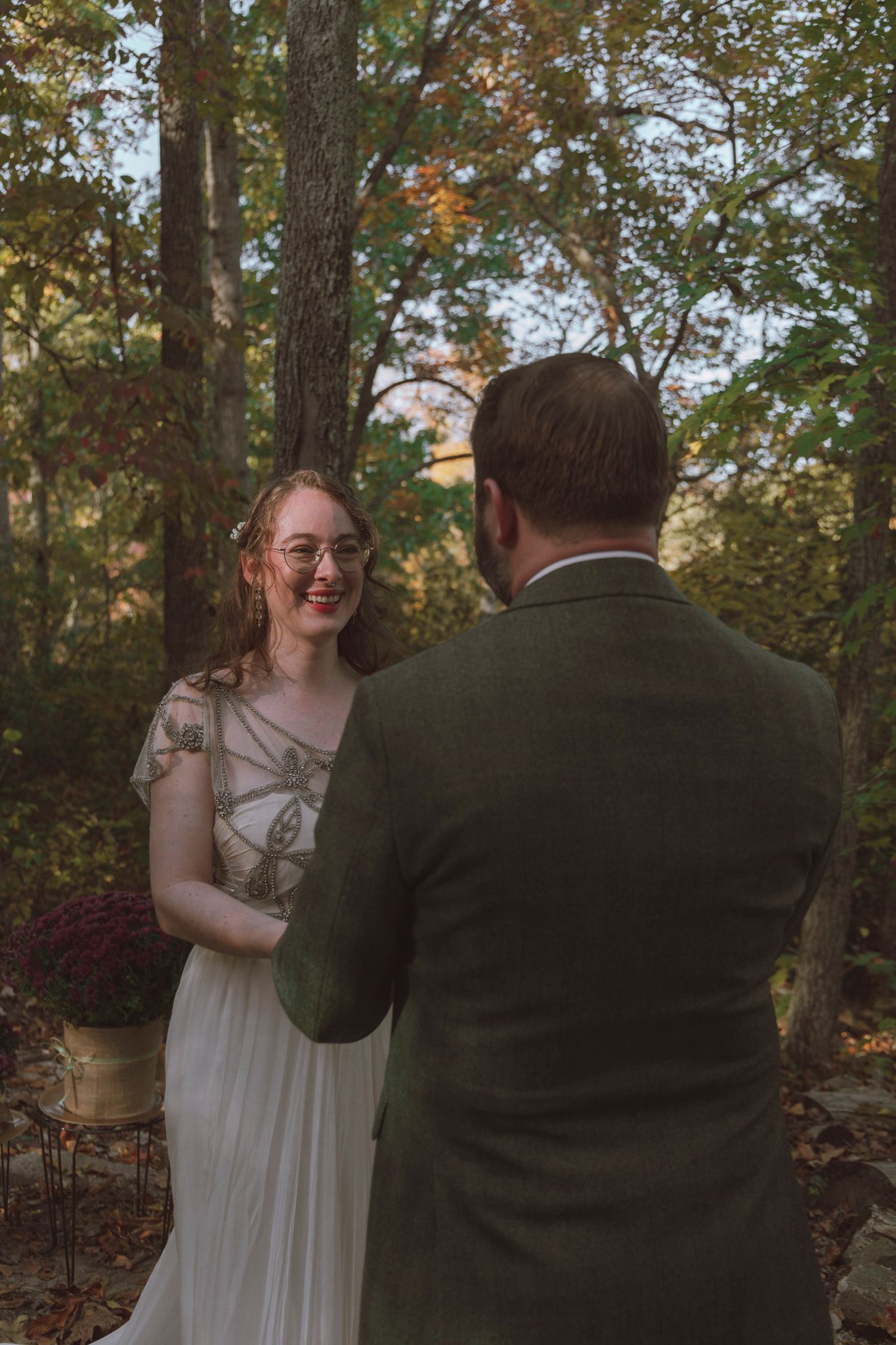 A couple shares a candid moment in a wooded outdoor setting during autumn with trees and foliage in the background.