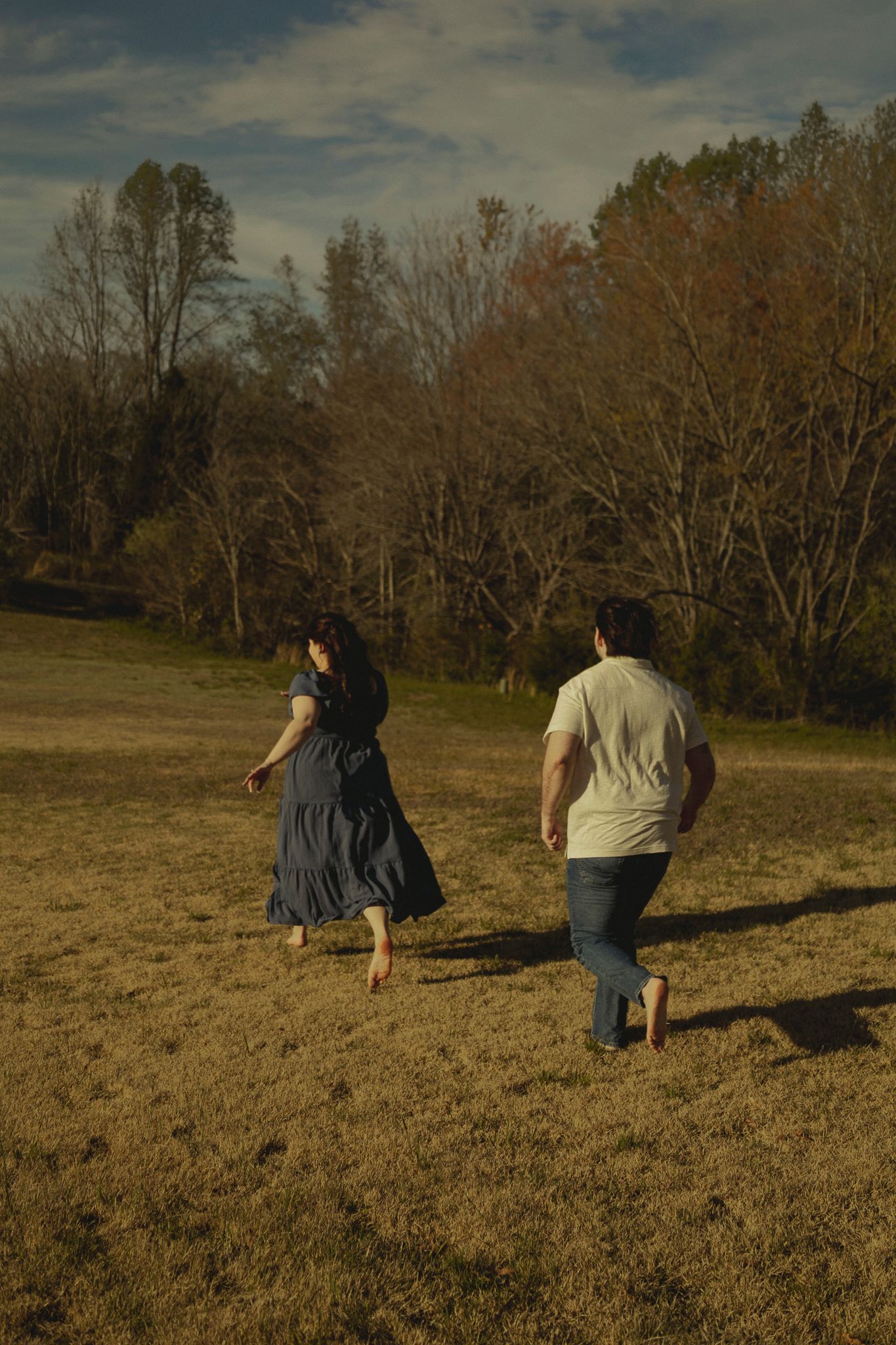 Couple running barefoot through golden field toward autumn trees at sunset.