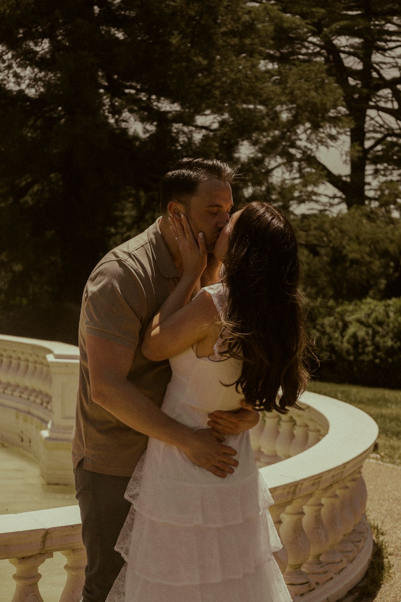Couple sharing a romantic kiss on a sunlit terrace with white balustrade railing and lush green trees in background.
