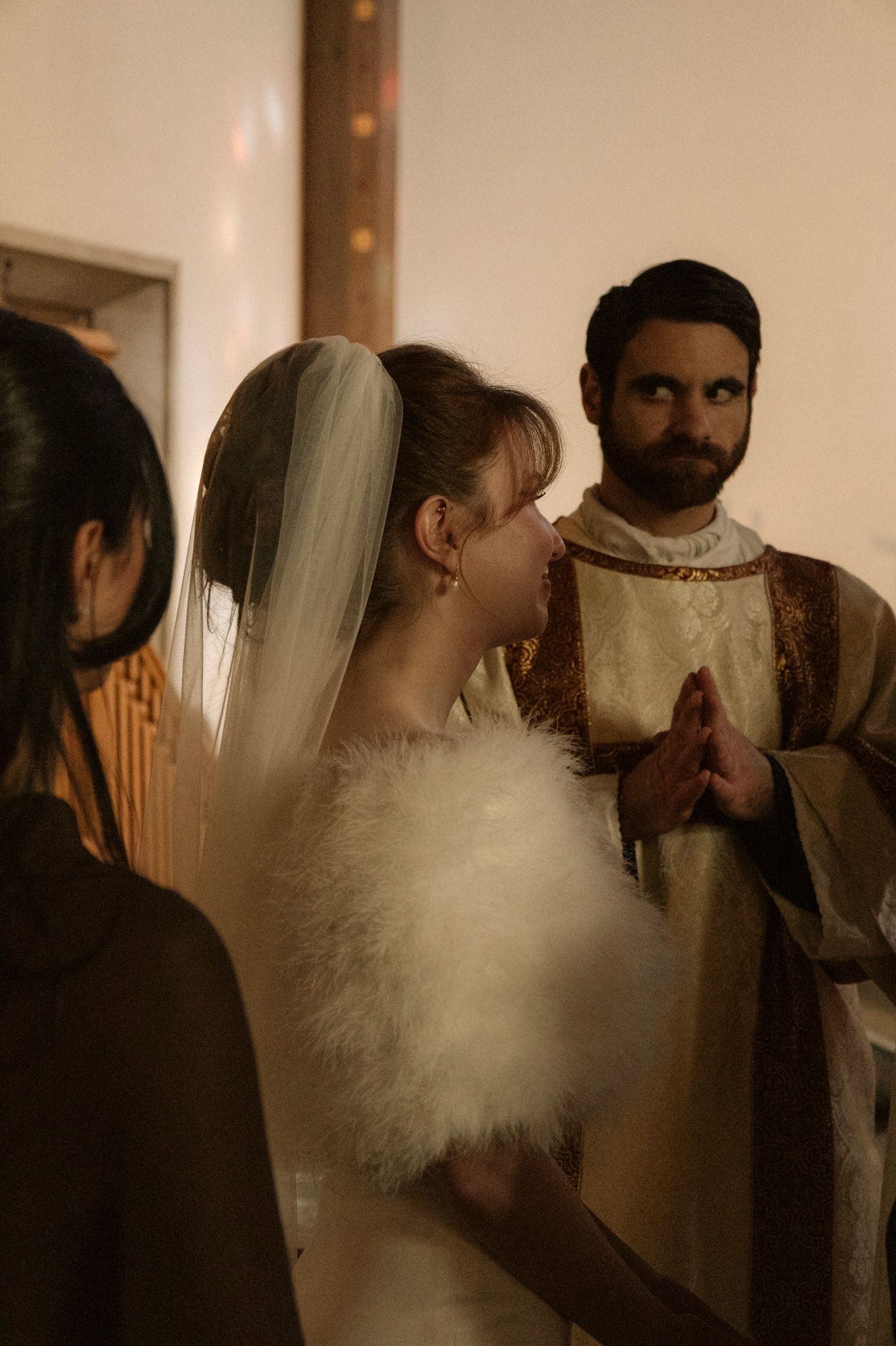 Bride in white gown with fur wrap and veil speaking with a bearded priest in golden vestments during a Catholic wedding.
