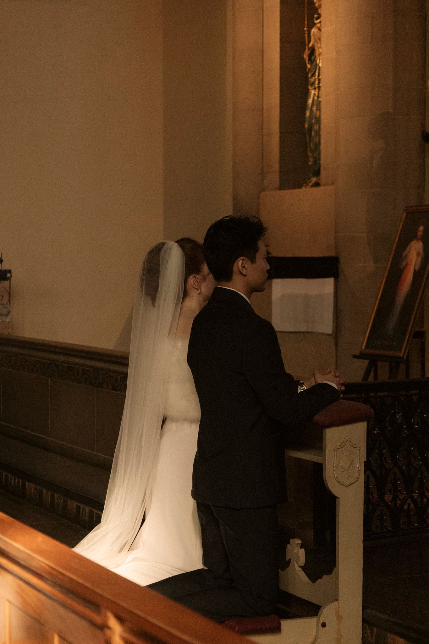 Bride and groom kneeling together in prayer at a wooden pew beside a religious painting during Catholic wedding ceremony.