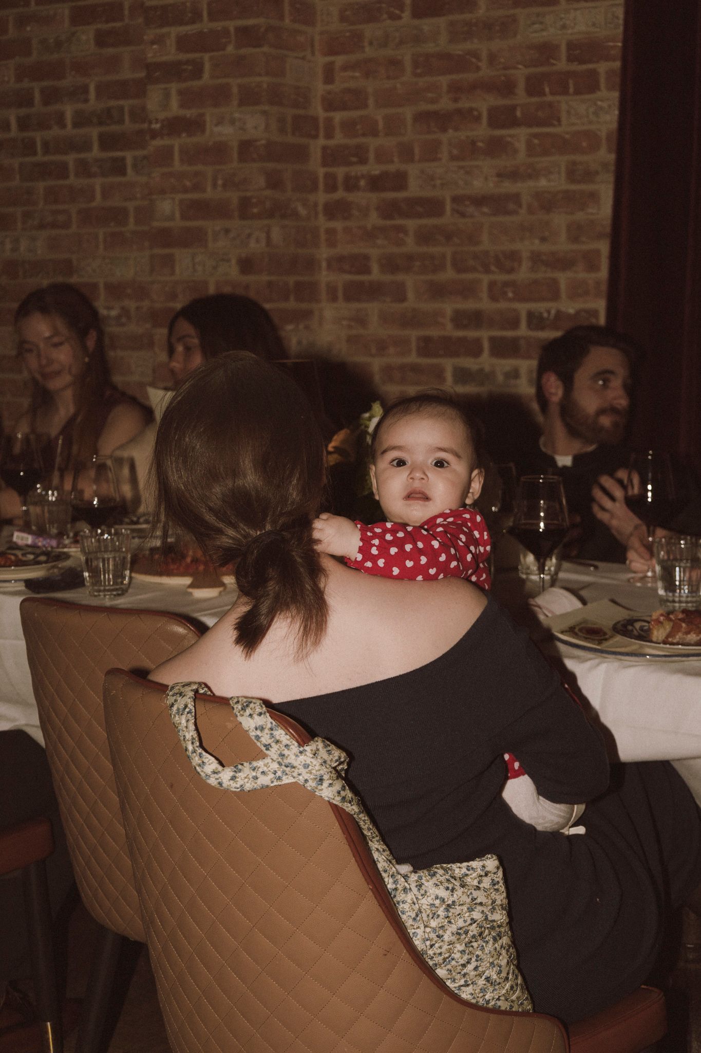 Mother holding baby in red polka dot outfit at a warm, dimly lit restaurant dinner event.