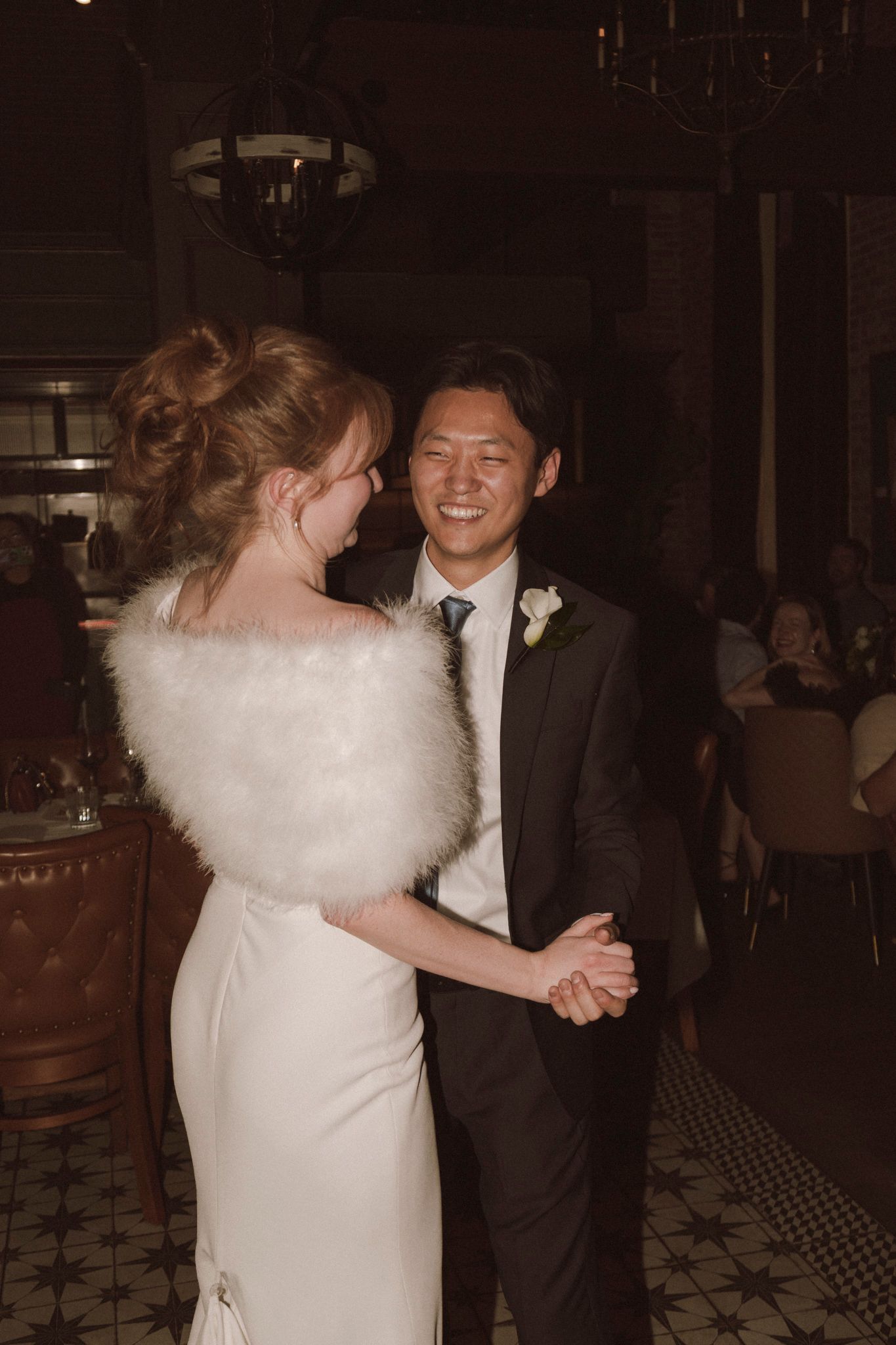 Bride in white gown with feather wrap shares first dance with groom in dark suit at wedding reception.