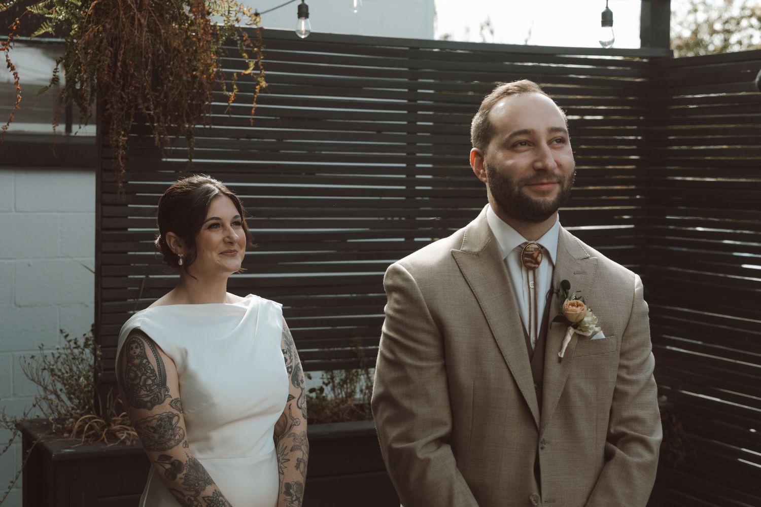 Newlyweds share intimate moment against modern slatted wall during outdoor wedding celebration.